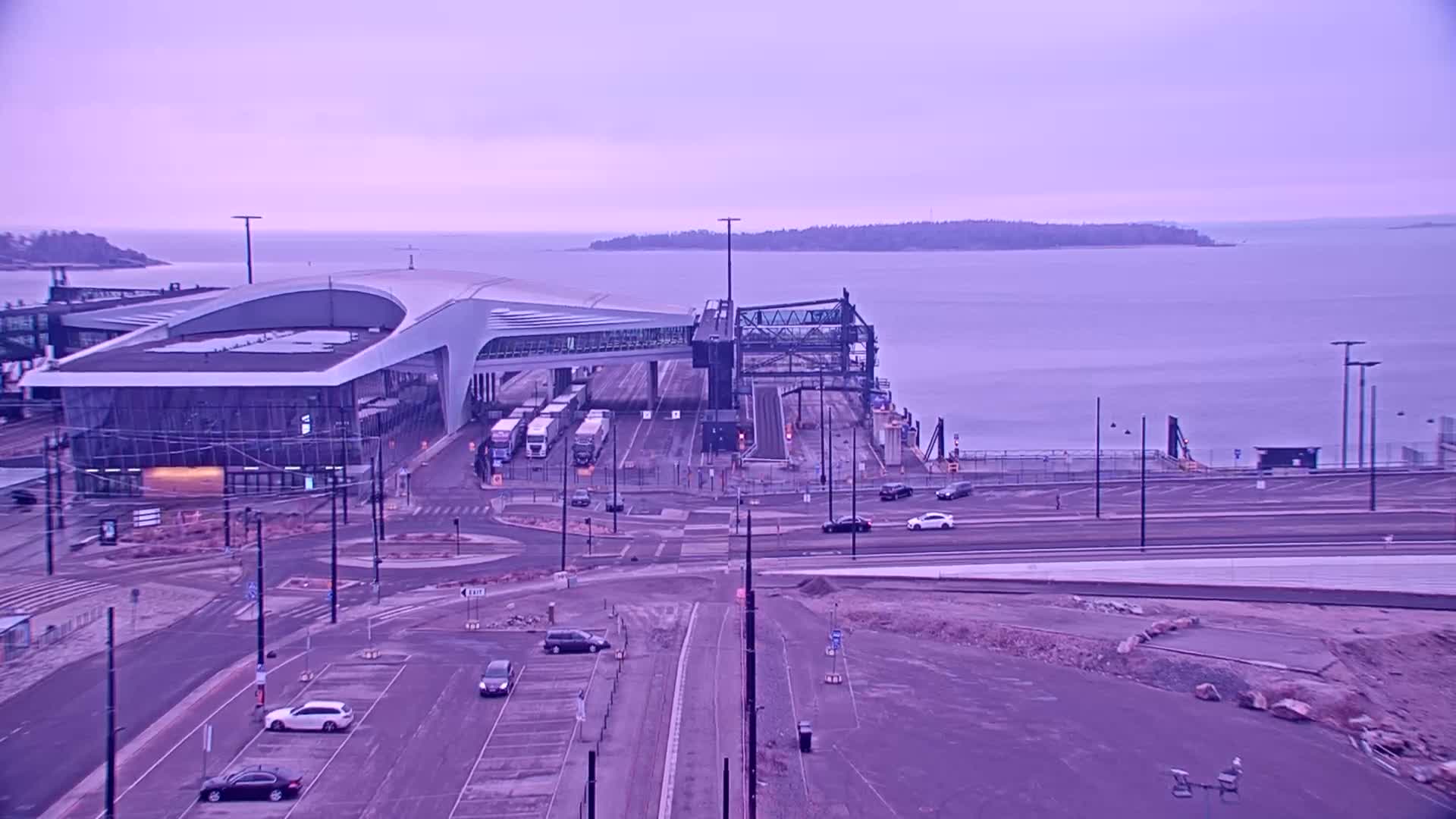 A modern ferry terminal with several vehicles and trucks is situated on the waterfront under a pale purple sky.