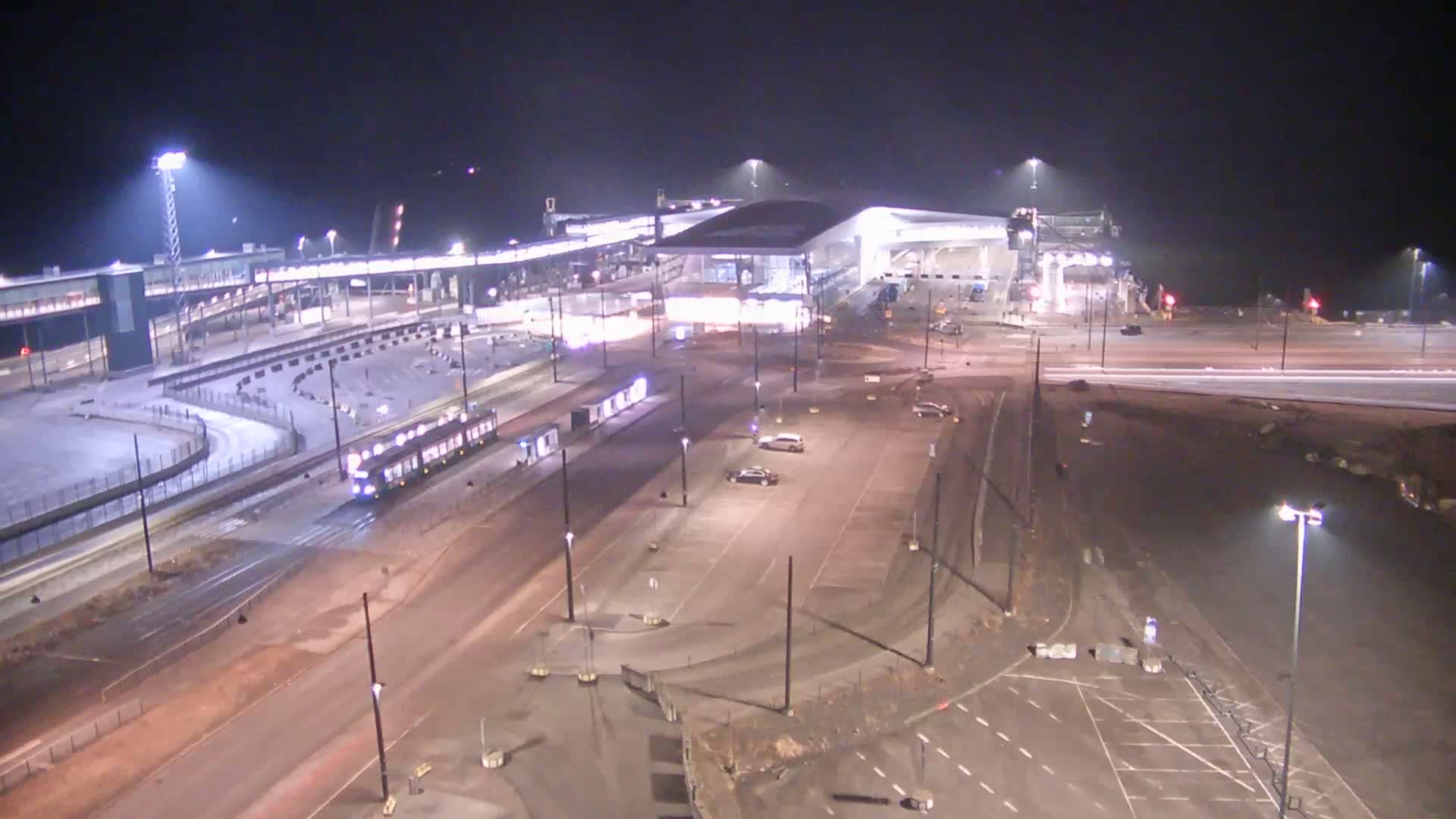 A nighttime, brightly lit view of a transportation hub with a tram traveling along a track, several cars parked in a lot, and a large modern building.