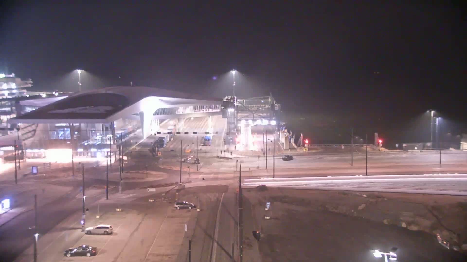 A nighttime, aerial view of a modern ferry terminal with a few cars in the parking lot and surrounding roads, under hazy or foggy conditions.