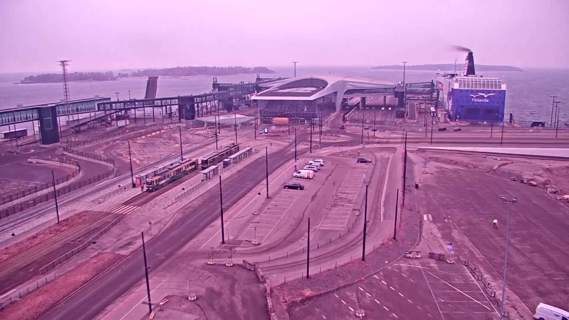 An aerial view of a ferry terminal with a ferry docked, a tram, cars in a parking lot, and ongoing construction under an overcast sky.
