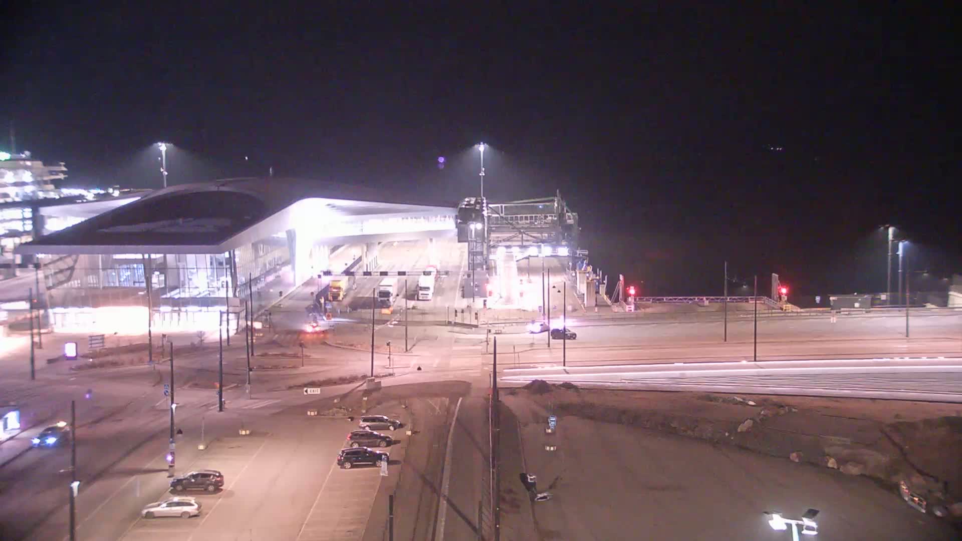 A brightly lit ferry terminal at night with several cars in the parking lot and a few vehicles near the ferry loading area.