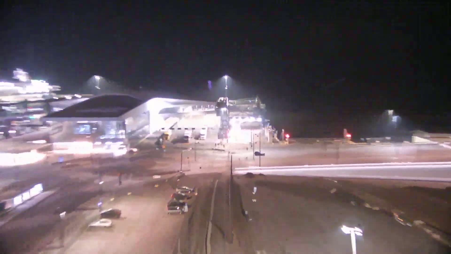 A nighttime, high-angle view shows a large, brightly lit building near a roadway intersection with several vehicles and streetlights visible.