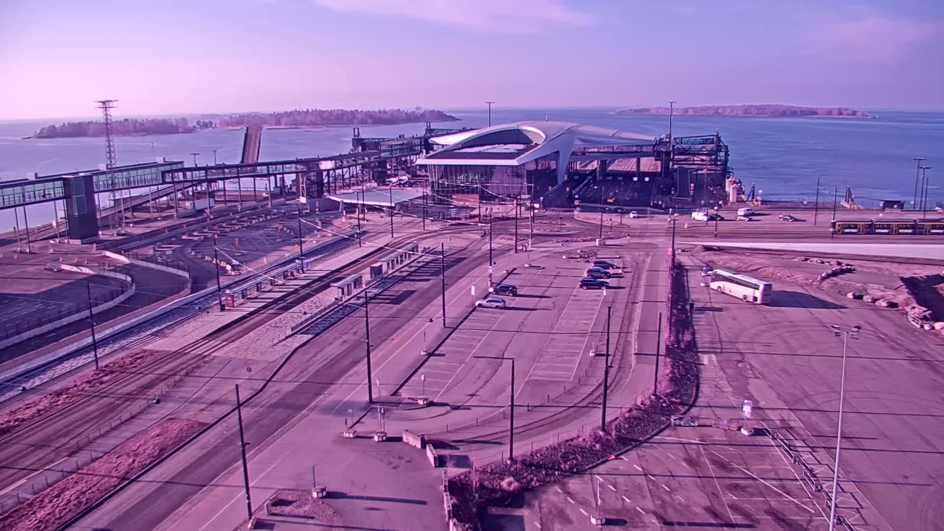 An aerial view shows a ferry terminal with a modern terminal building,  parking lots, and transit infrastructure, under a clear, bright sky.