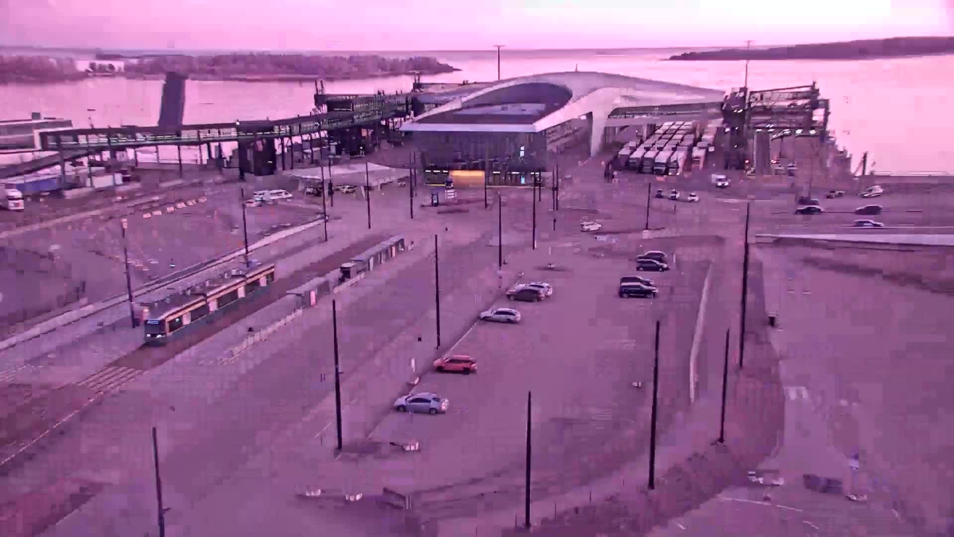 An aerial view shows a ferry terminal with a tram, several cars in a parking lot, and numerous trucks, all under a dusky pink sky.