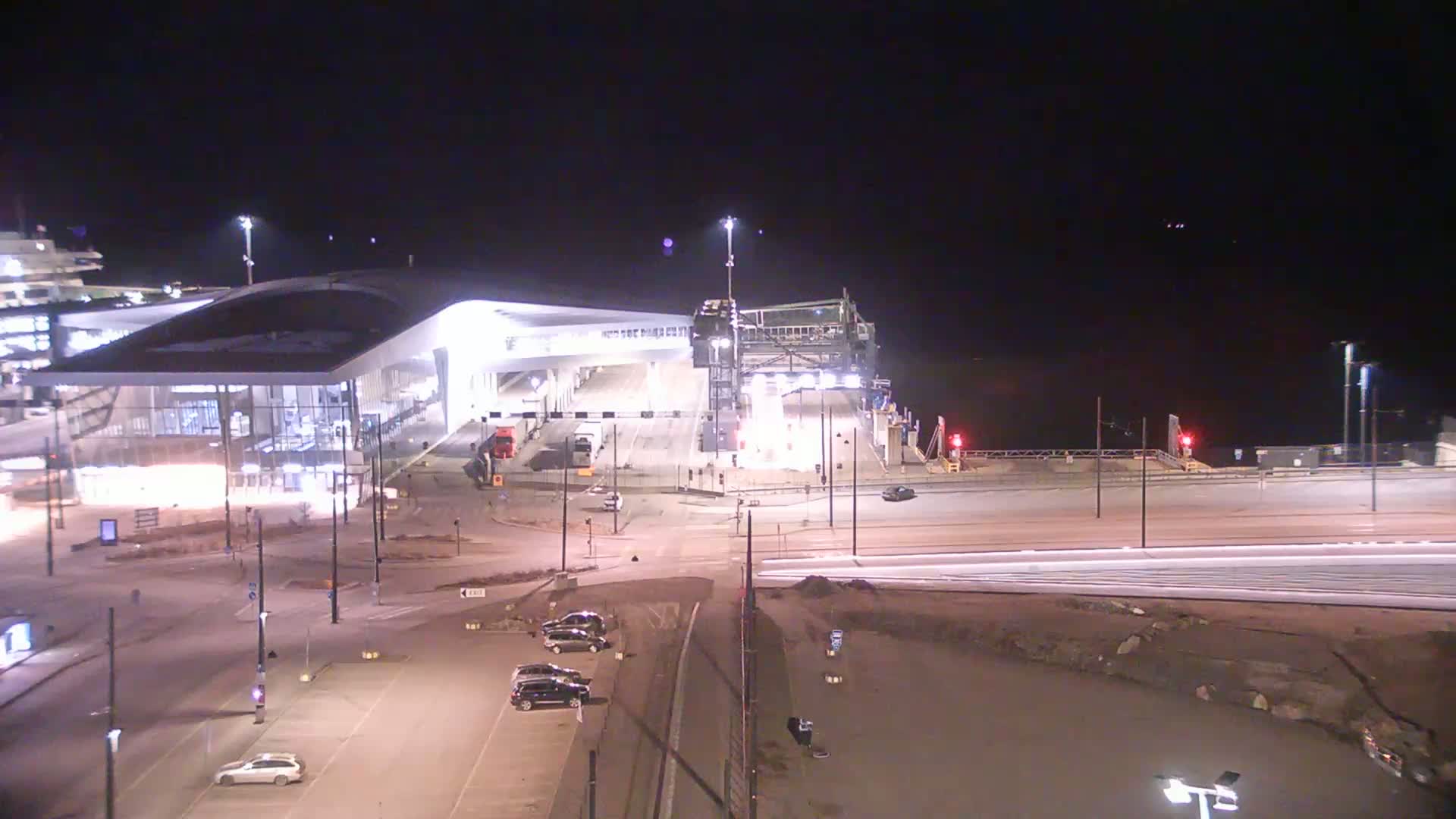 A brightly lit ferry terminal at night, with a few cars parked nearby and a dark body of water in the background.