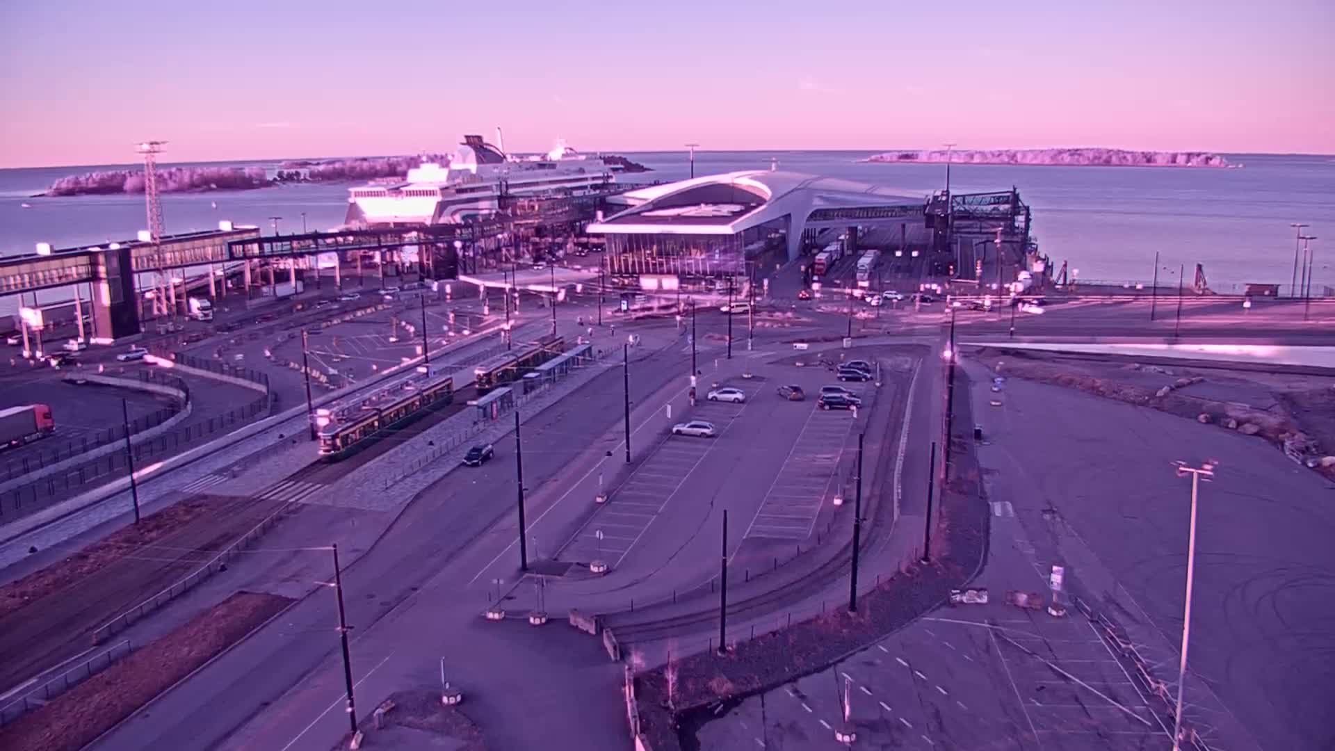 A ferry terminal with a tram, cars in a parking lot, and a calm body of water under a pinkish-purple twilight sky.