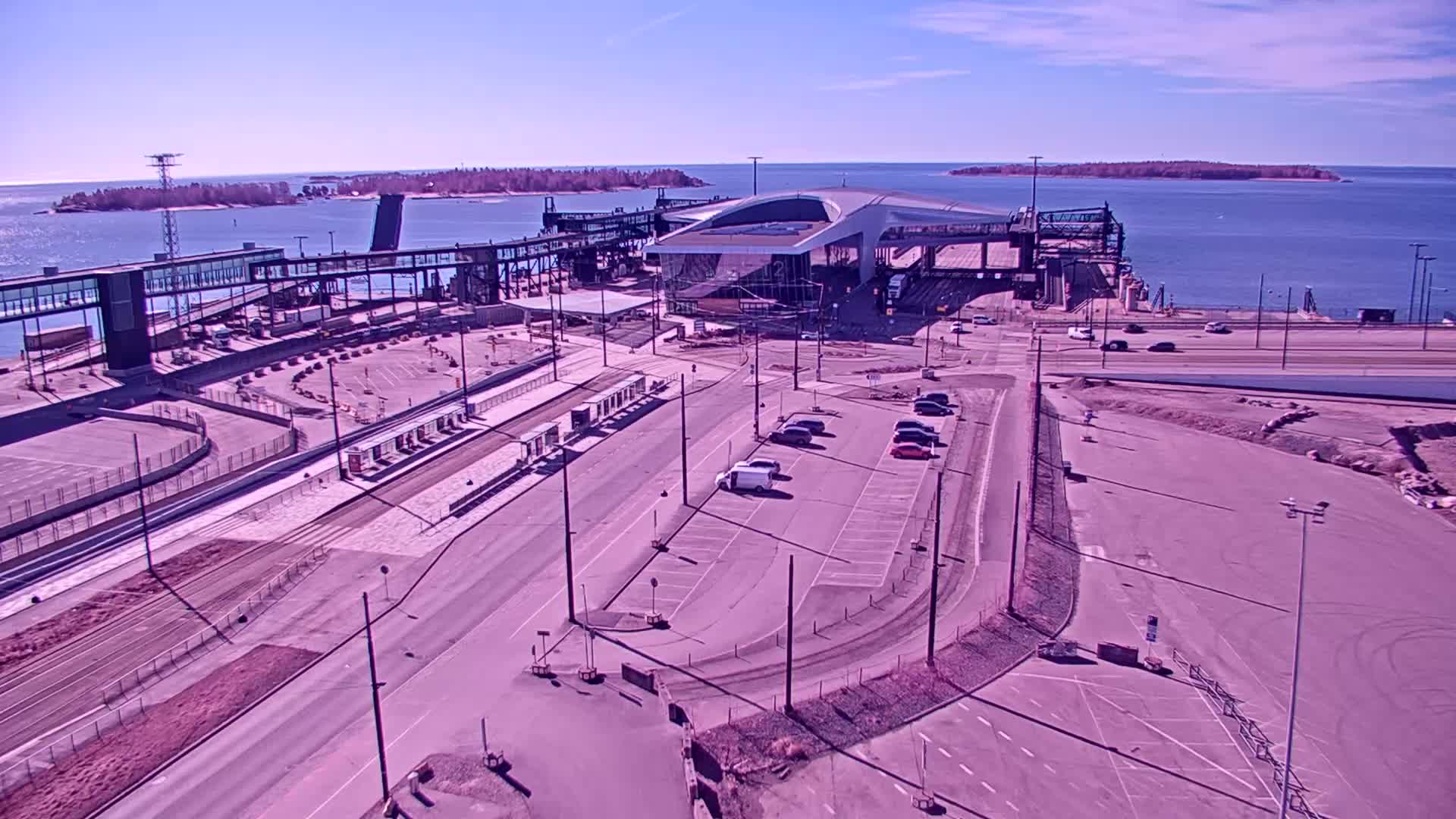 A modern ferry terminal with a curved roof and surrounding parking areas and roadways is situated on a waterfront on a sunny day.
