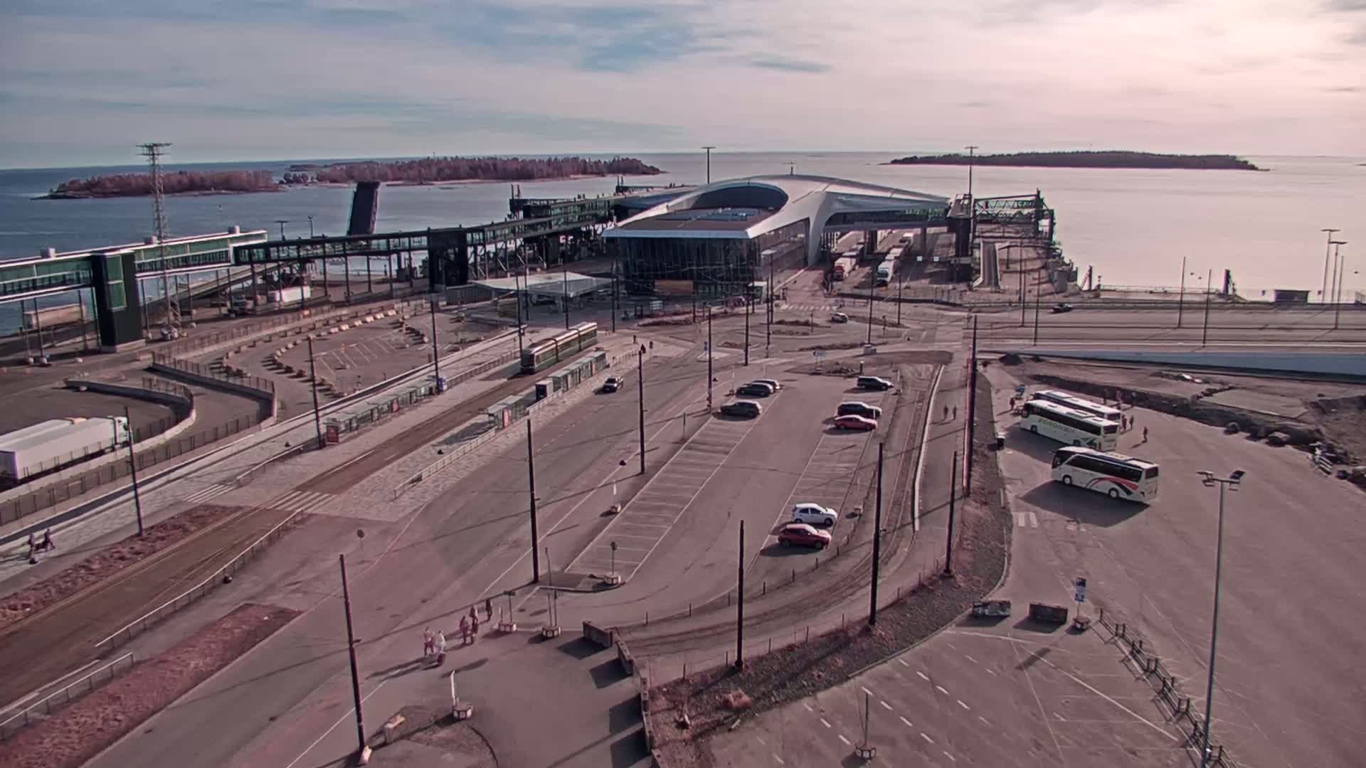 An aerial view of a ferry terminal with several cars and buses parked nearby, under a partly cloudy sky.