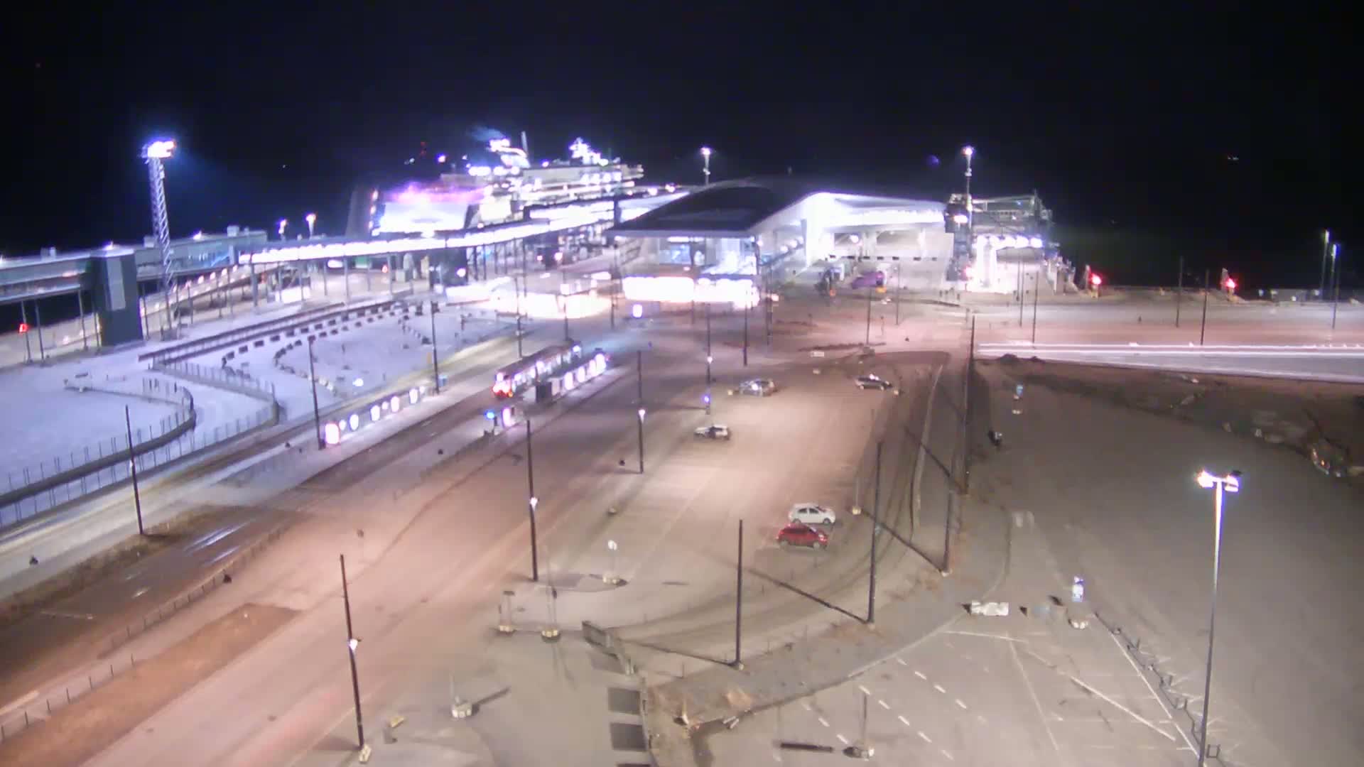 A nighttime aerial view shows a cruise ship docked at a terminal, with a tram stopped near a mostly empty parking lot and roadway.