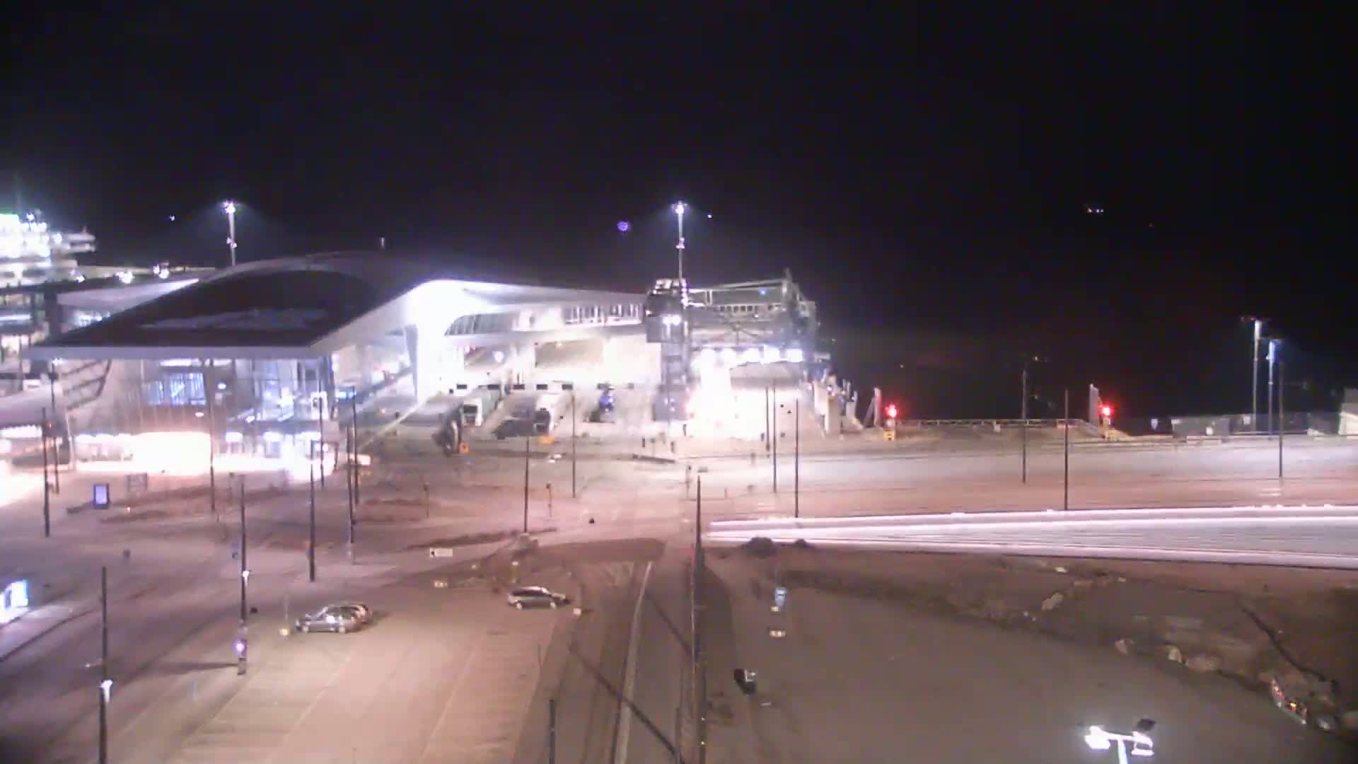 A brightly lit ferry terminal at night with a few cars in the parking lot and light streaks from moving vehicles on the road.