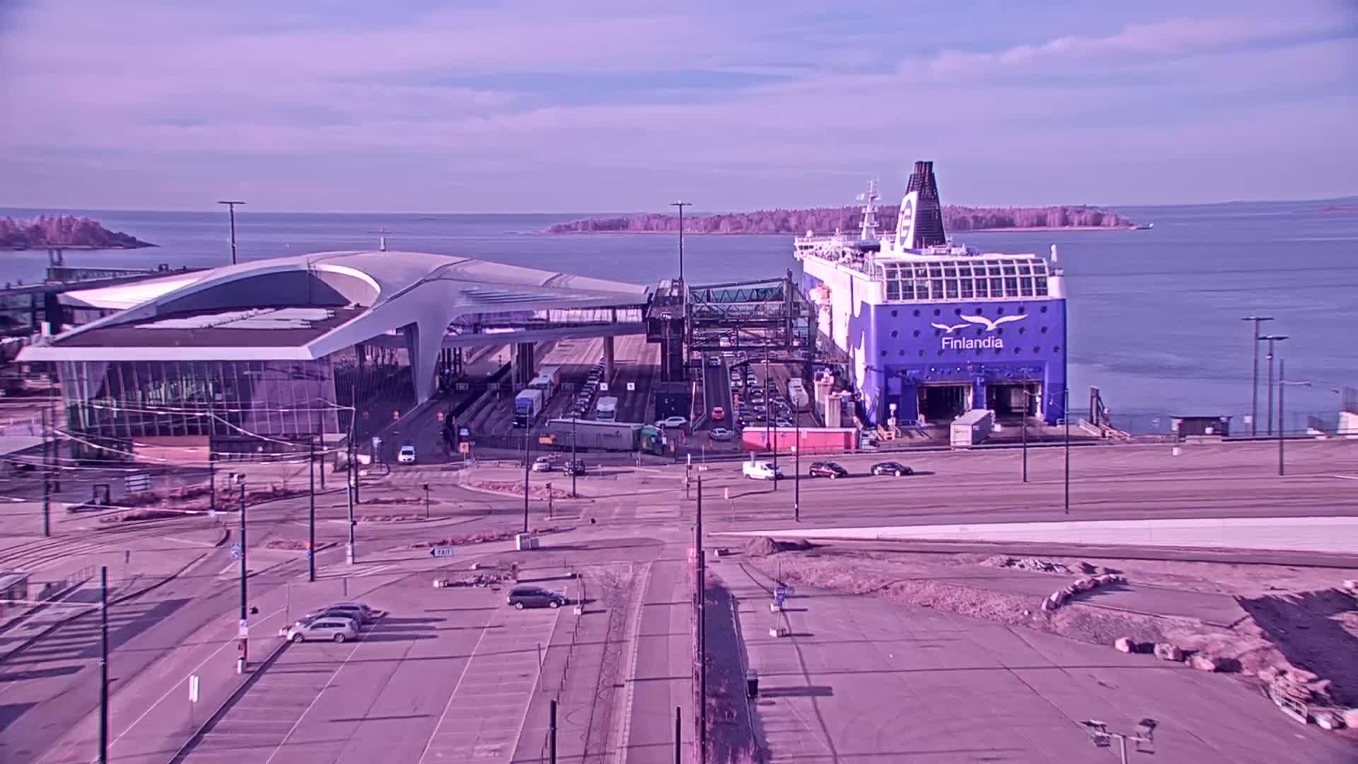 A large ferry is docked at a terminal, with cars and trucks in the surrounding area under a clear, sunny sky.