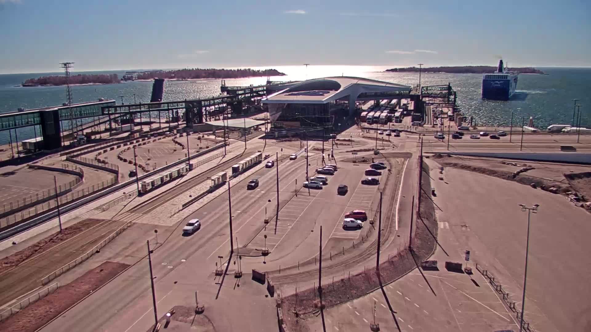 An aerial view of a ferry terminal on a sunny day shows a large ferry departing, along with a parking lot, roadways, and several buses near the terminal building.