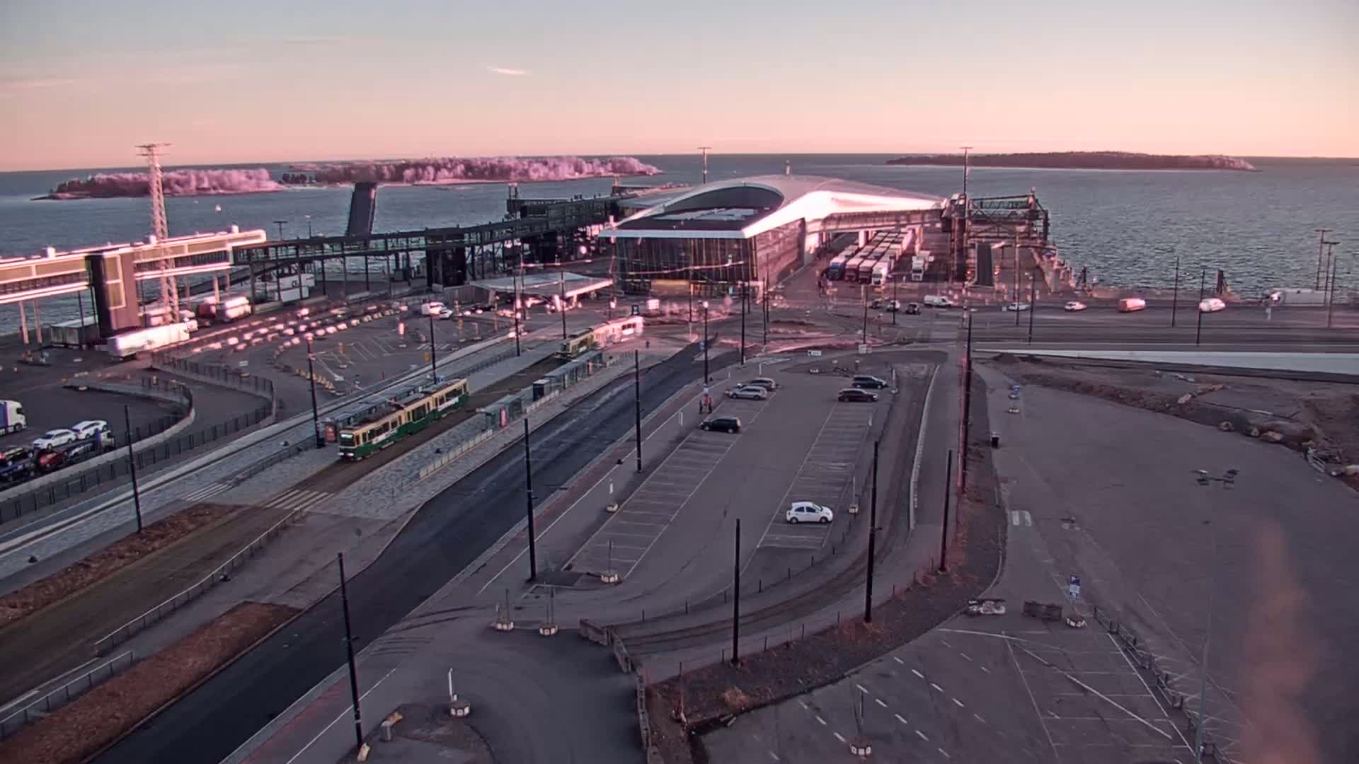 An aerial view of a ferry terminal at sunrise, with a tram on the tracks nearby, and several vehicles in parking lots.