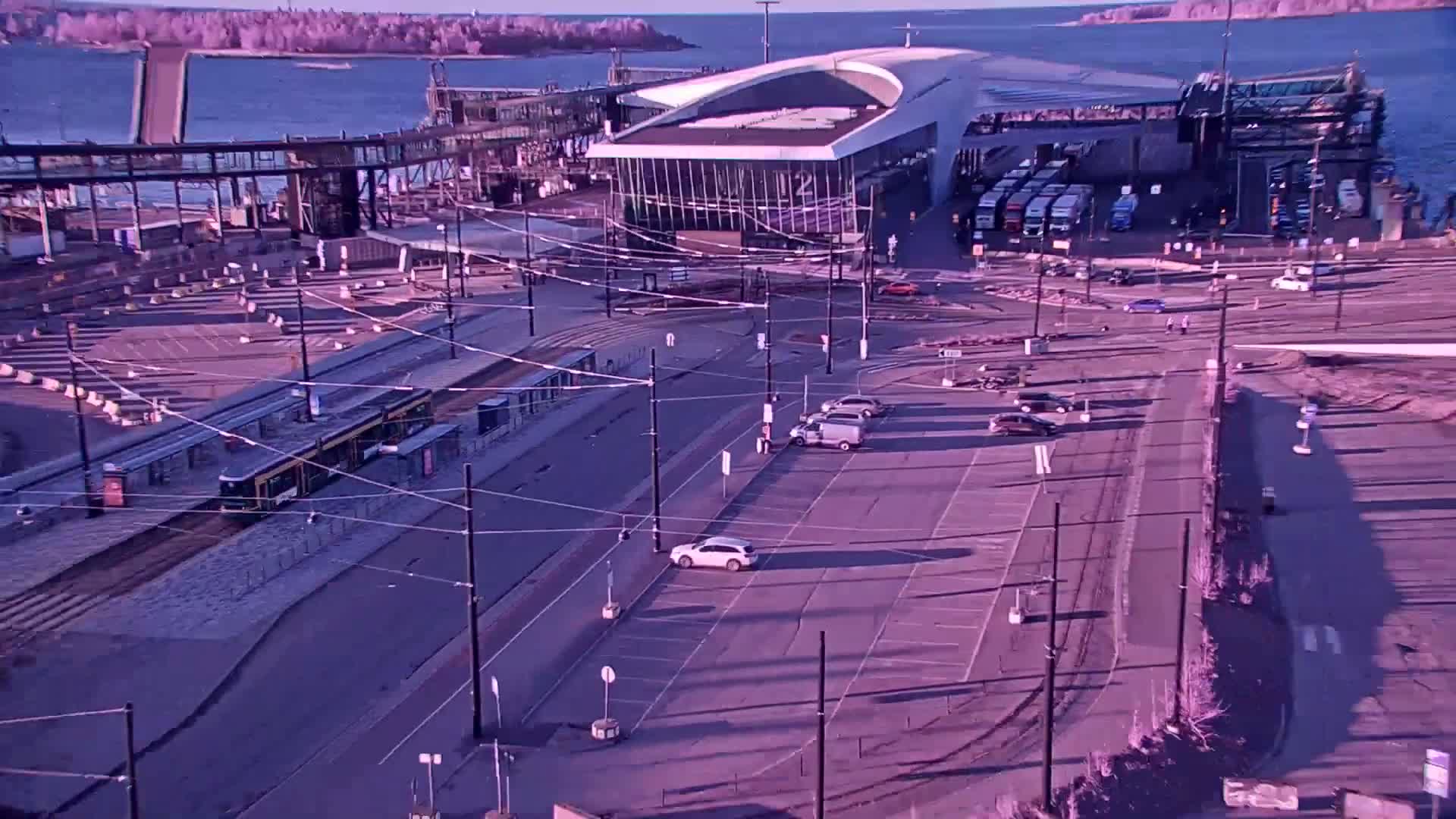 An aerial view shows a tram at a station near a ferry terminal and parking lot, under a clear, bright sky over calm water.