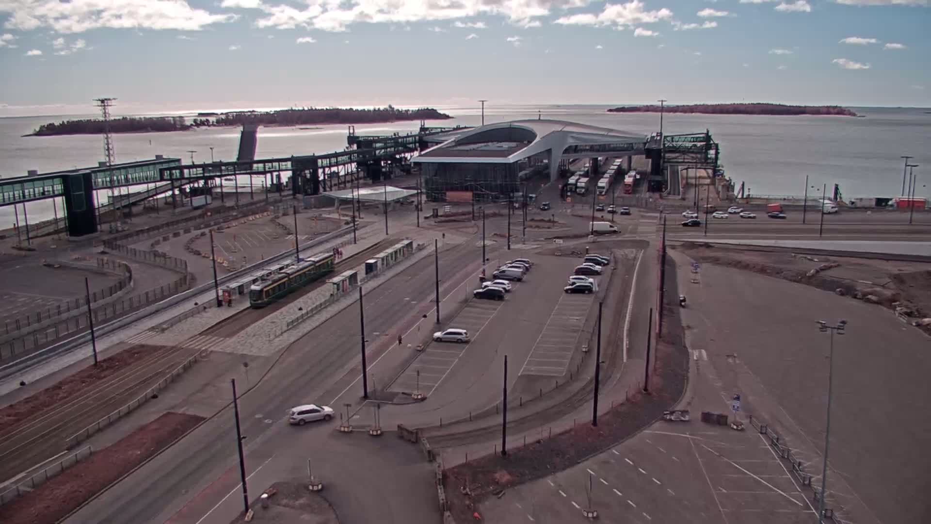 An aerial view shows a ferry terminal with a tram, cars in a parking lot, and buses, under a partly cloudy sky.