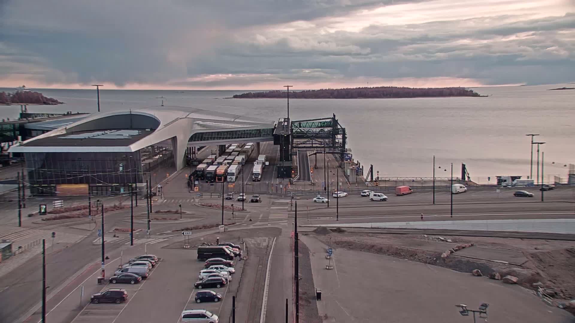 A ferry terminal with several vehicles parked nearby and trucks docked under a covered walkway, overlooking a calm body of water under an overcast sky.