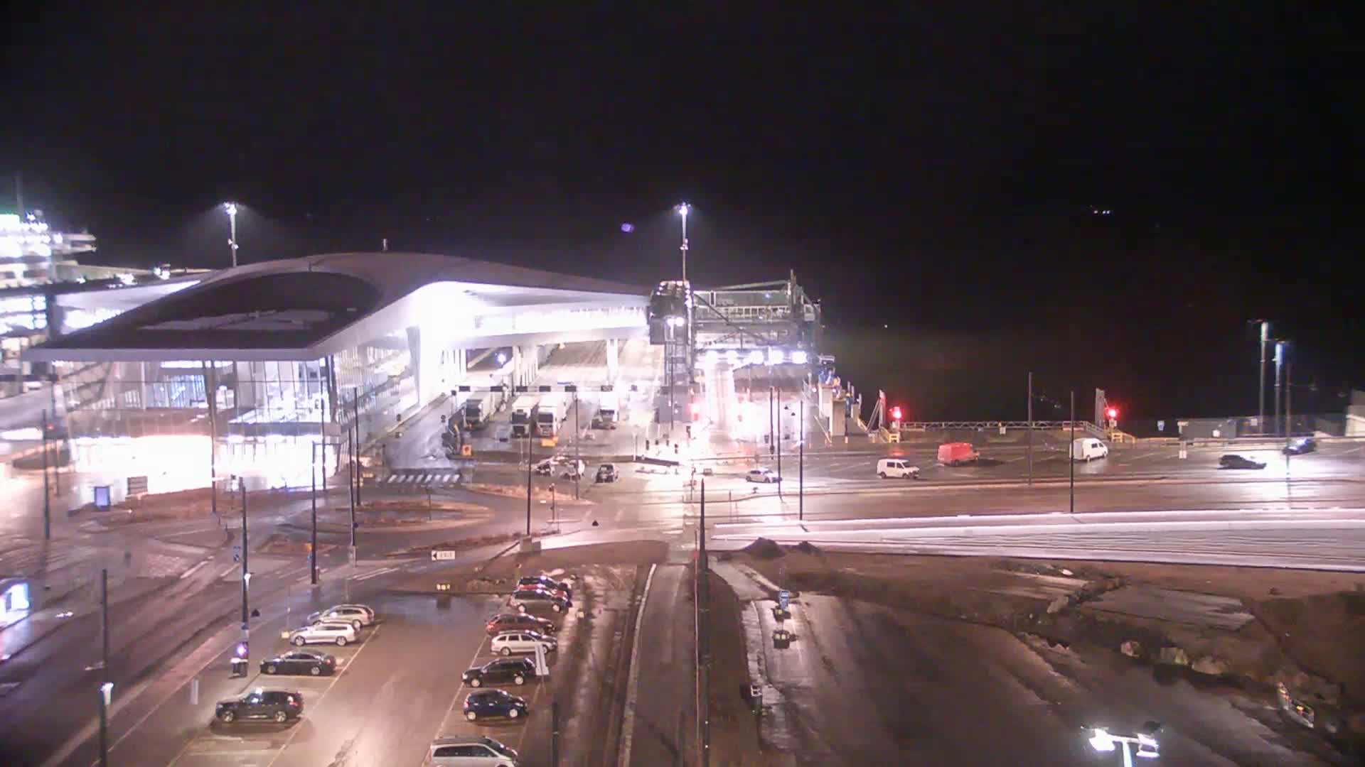 A brightly lit ferry terminal at night, with vehicles parked nearby and a ferry docked at the terminal under a dark sky.