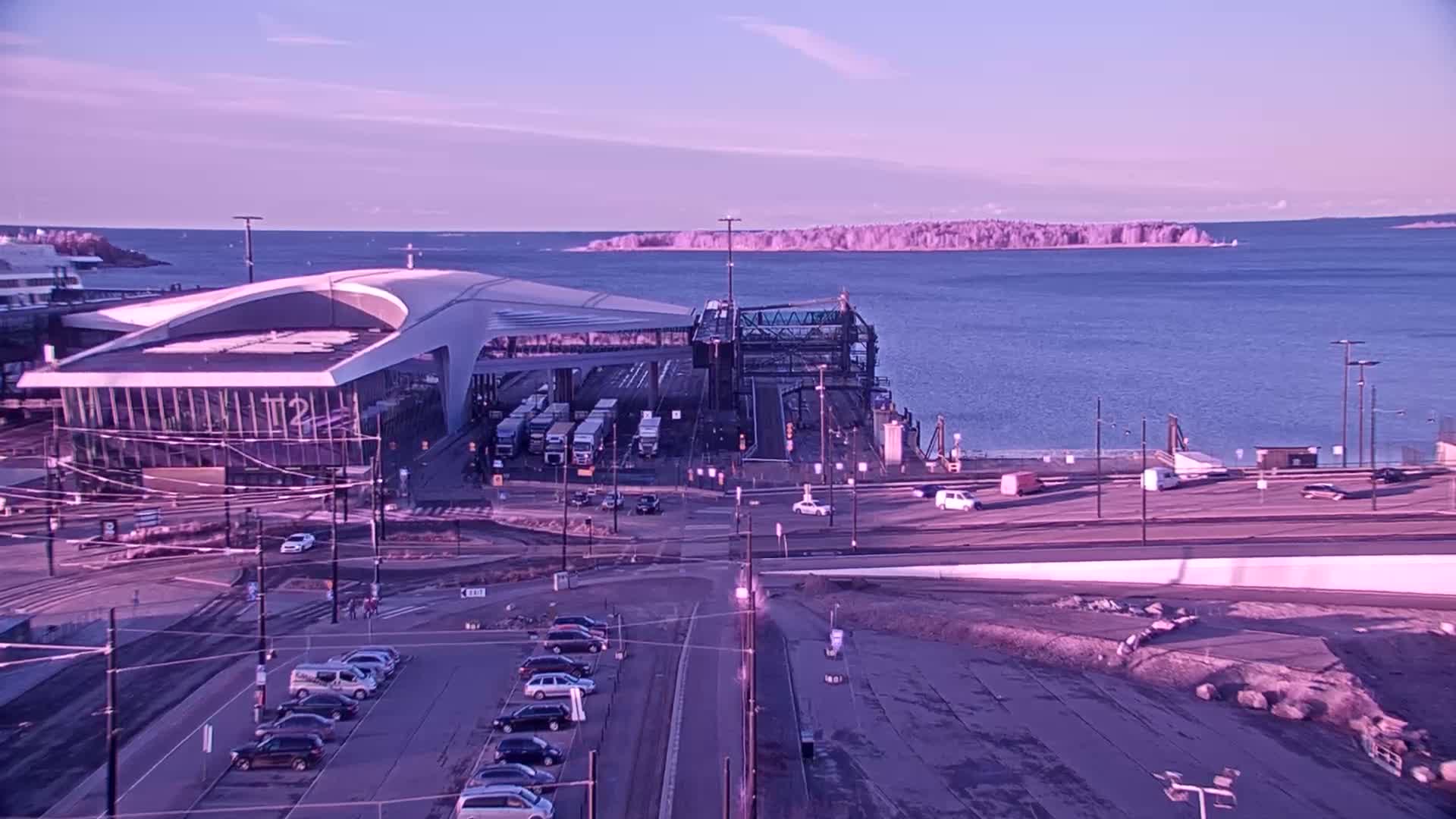 A ferry terminal with several vehicles parked nearby, situated on a body of water under a clear, purplish-hued sky.