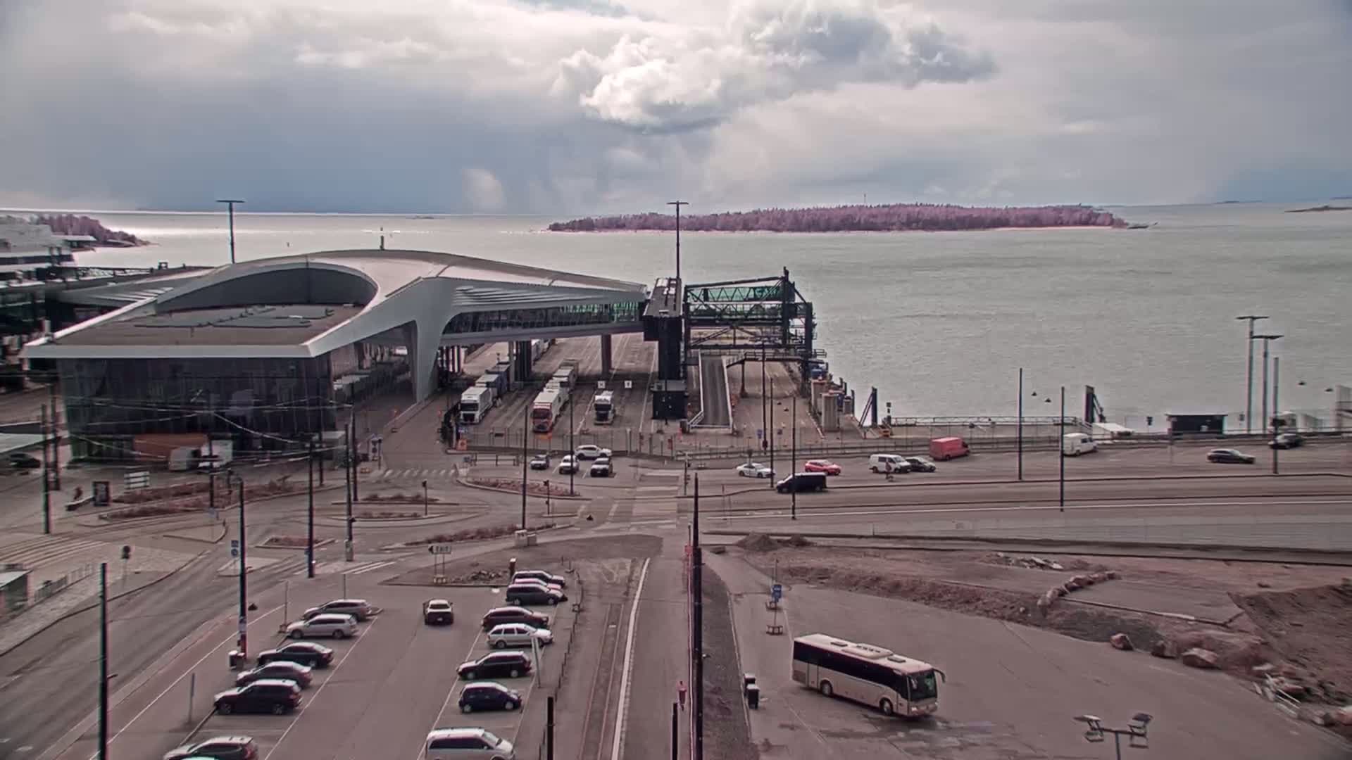 A ferry terminal with a modern building and several vehicles is shown, under an overcast sky, next to a body of water with an island in the distance.