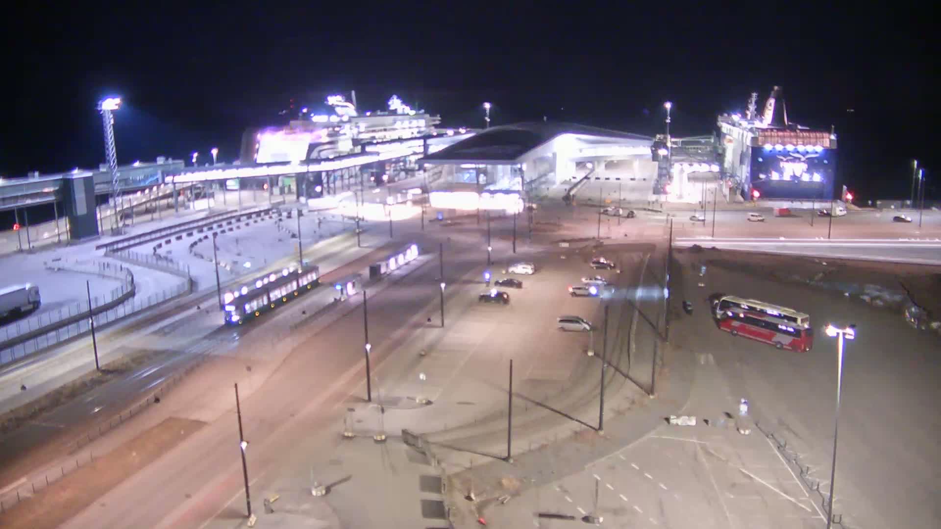 A nighttime aerial view of a ferry terminal with a large ferry docked, several vehicles in a parking lot, and a tram on the road.
