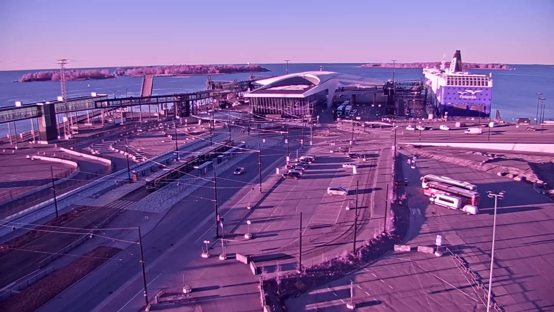 A ferry is docked at a terminal, with a tram, buses, and cars visible in a large parking lot under a clear, light purple sky.