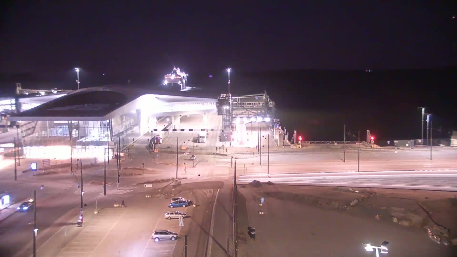 A ferry terminal at night, brightly lit, with a ferry docked and several cars in the parking lot.
