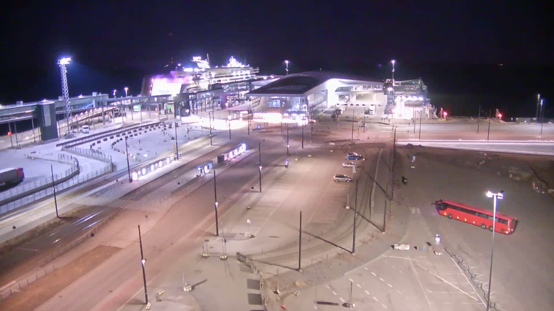 A large cruise ship is docked at a terminal at night, with a bus and several cars in a nearby parking lot under partly cloudy skies.