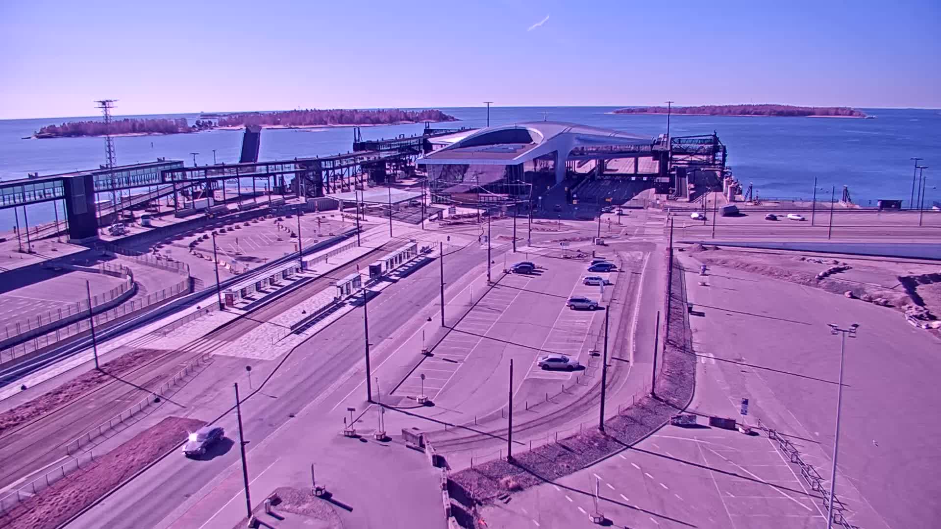 A ferry terminal with a modern building and parking areas is shown on a sunny day, next to calm blue water and several small islands in the distance.