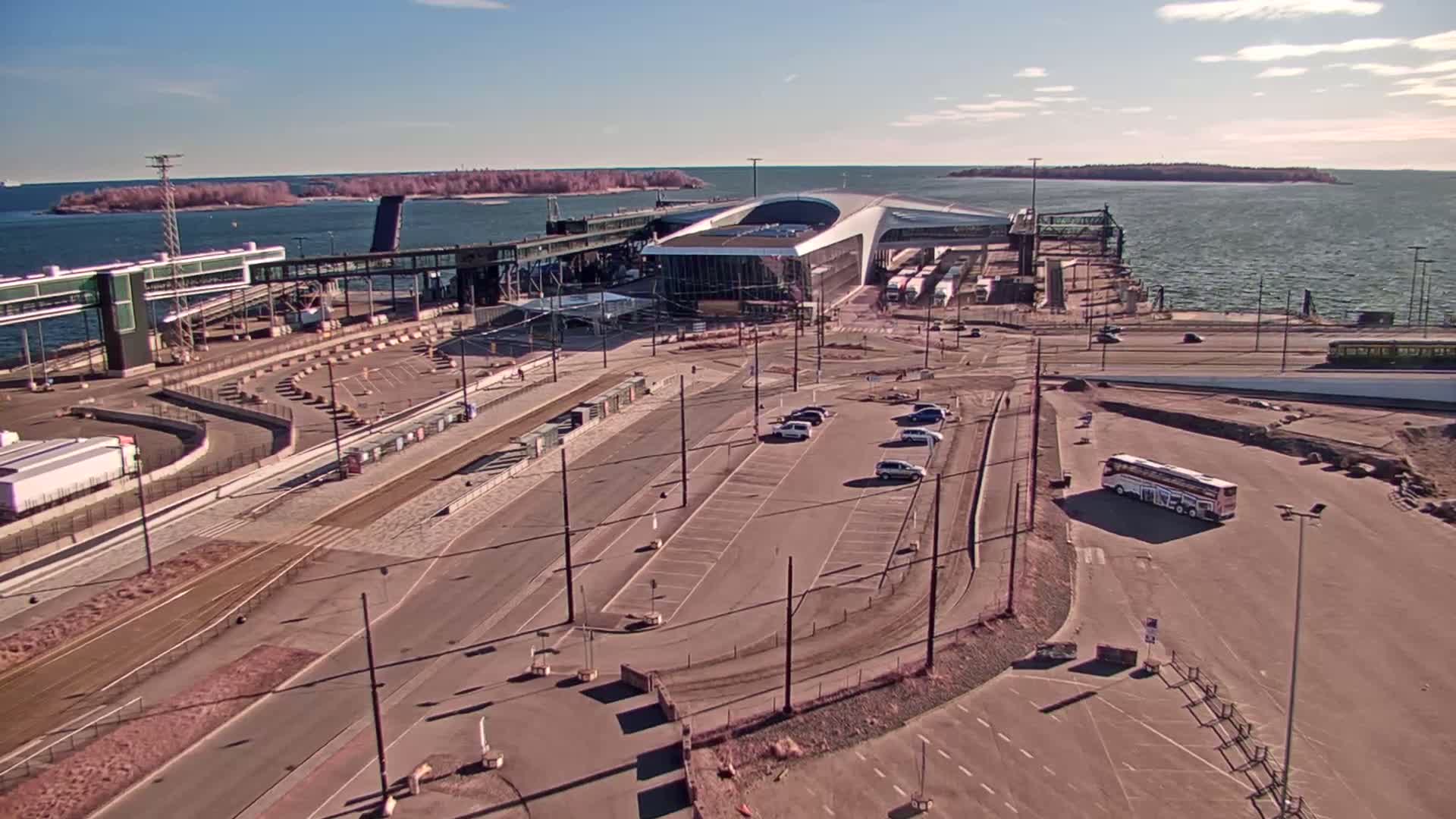 An aerial view shows a ferry terminal with a modern building, adjacent parking lots with a few cars and a bus, and a body of water with islands in the background under a sunny sky.