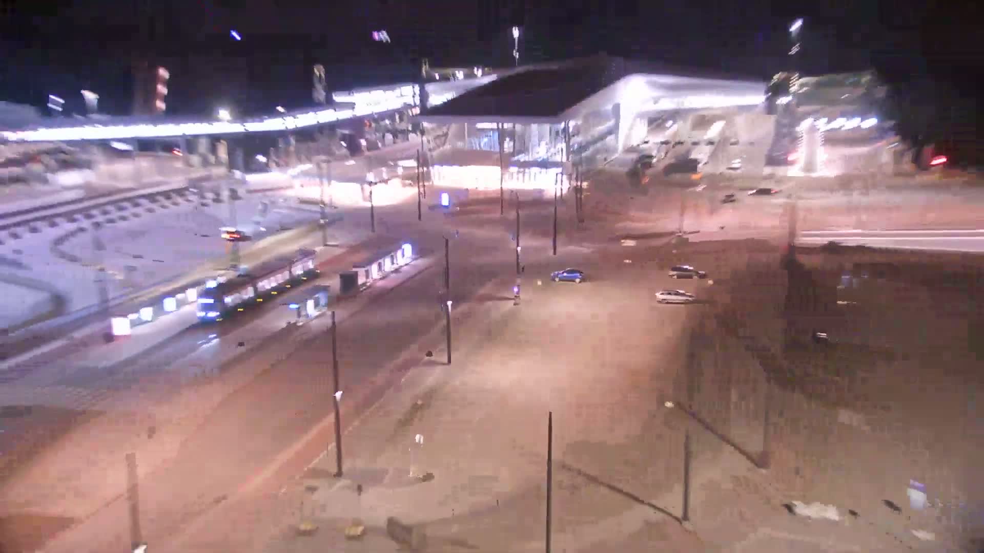 At night, a tram travels along a roadway near a large, modern building and a mostly empty parking lot.