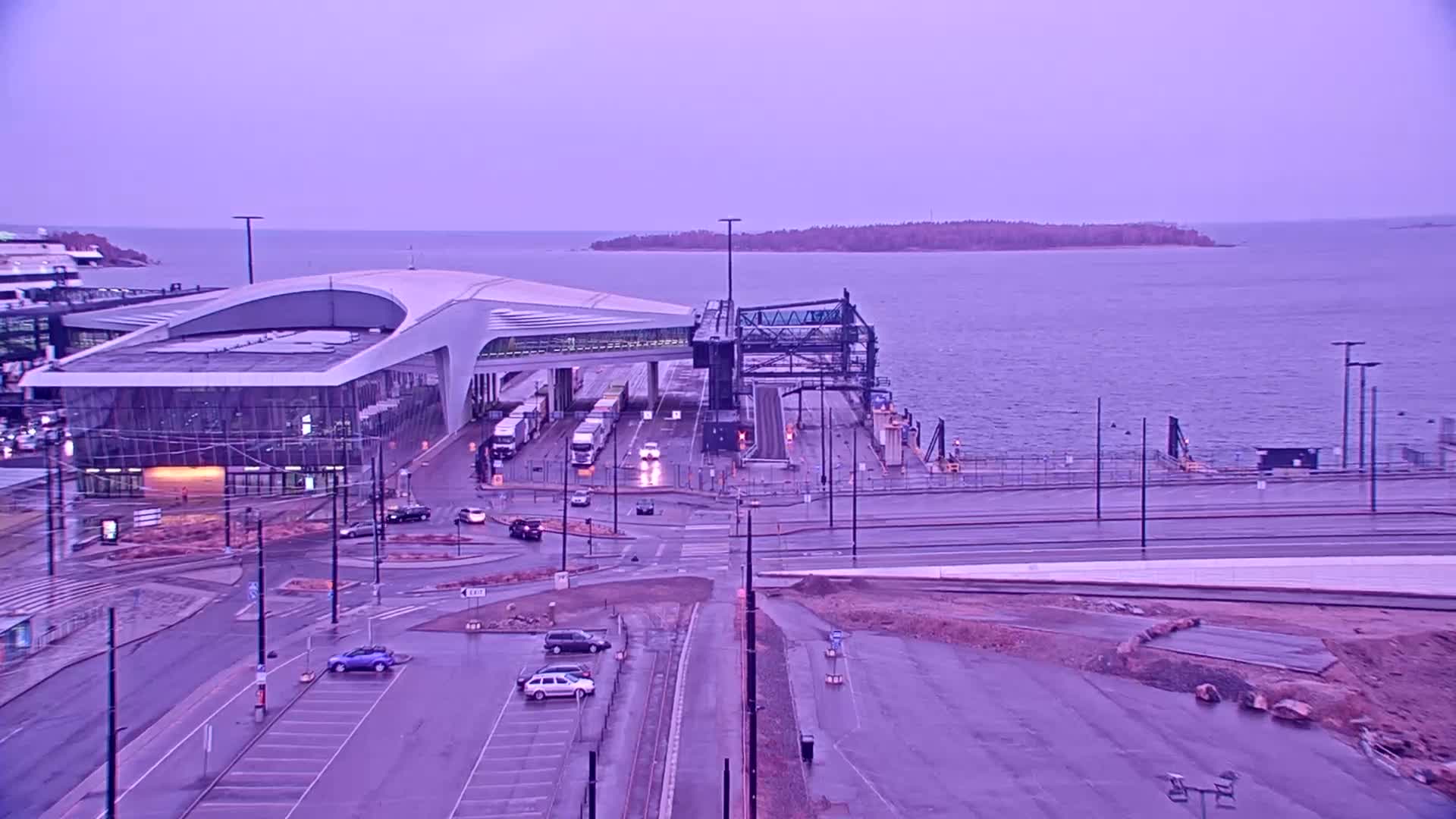 A modern ferry terminal with several vehicles parked nearby sits on a waterfront under a pale purple sky.