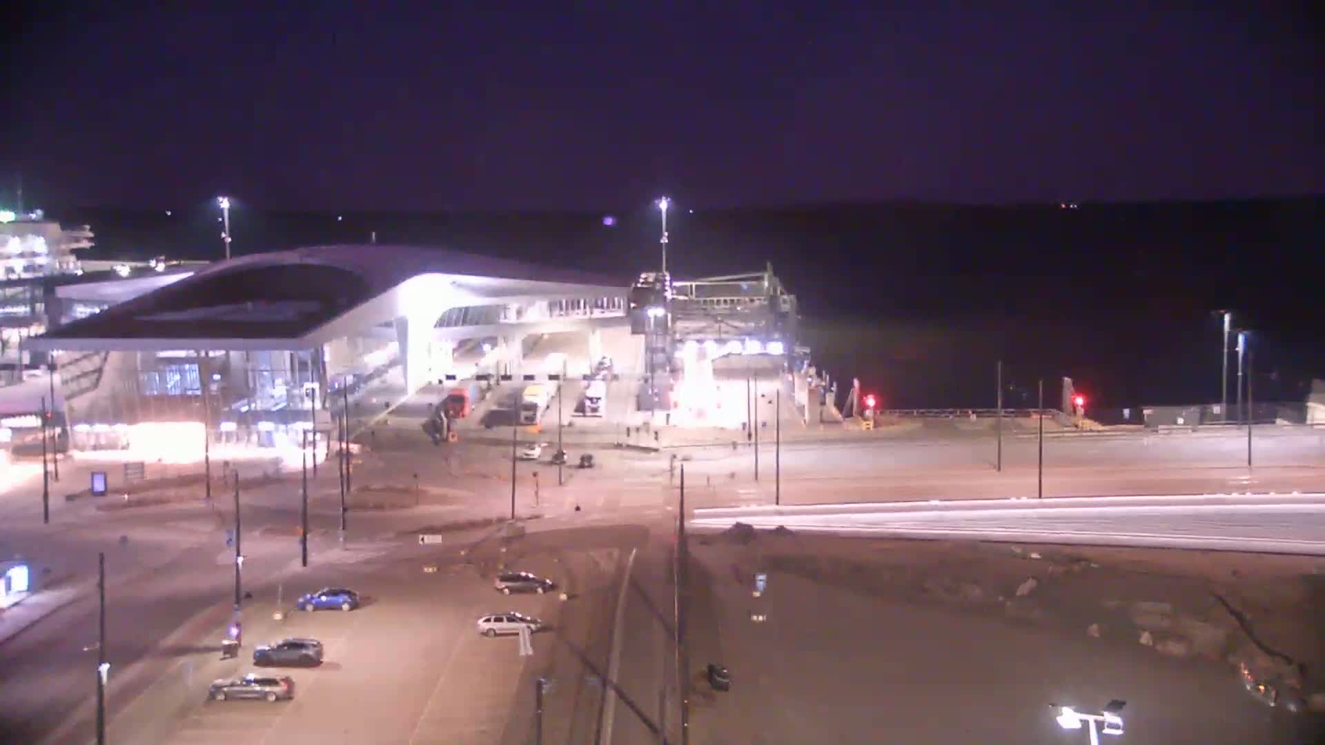 A ferry terminal at night, illuminated under a dark sky, with several cars parked in a nearby lot.