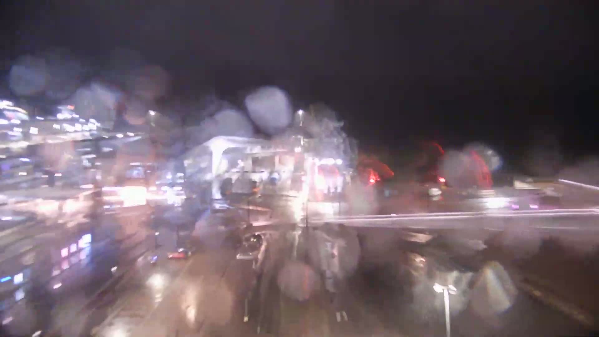 A rain-streaked view of a nighttime city street shows blurred traffic and buildings.