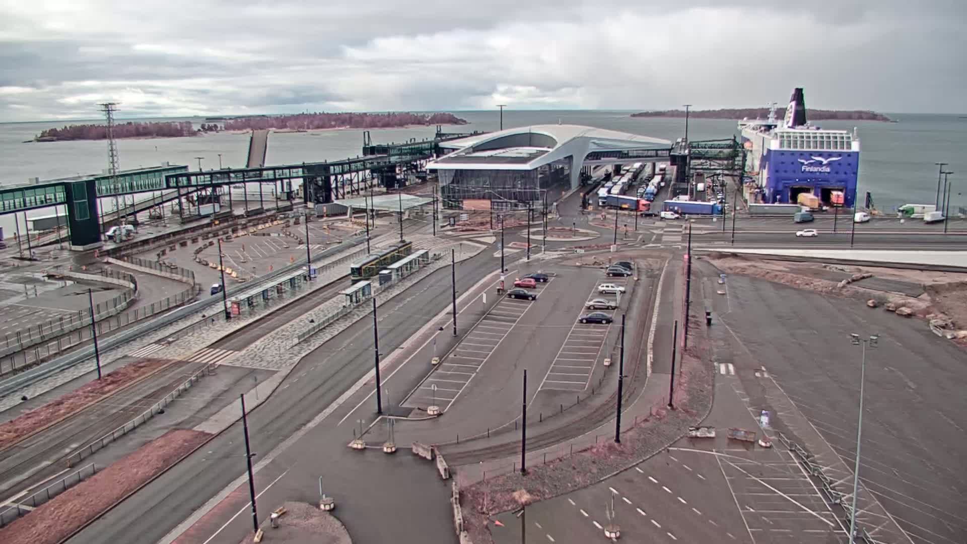 An aerial view of a ferry terminal on an overcast day, showing a large ferry docked, passenger walkways, parking lots, and a tram stop.