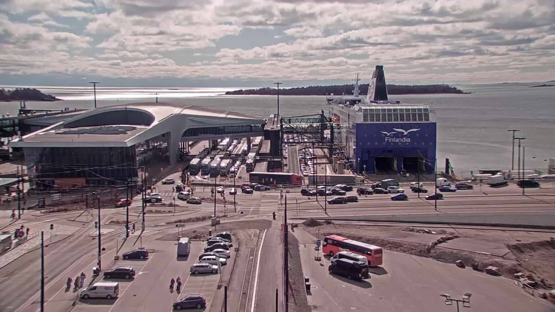 A large ferry is docked at a terminal, surrounded by vehicles and a modern terminal building under partly cloudy skies.