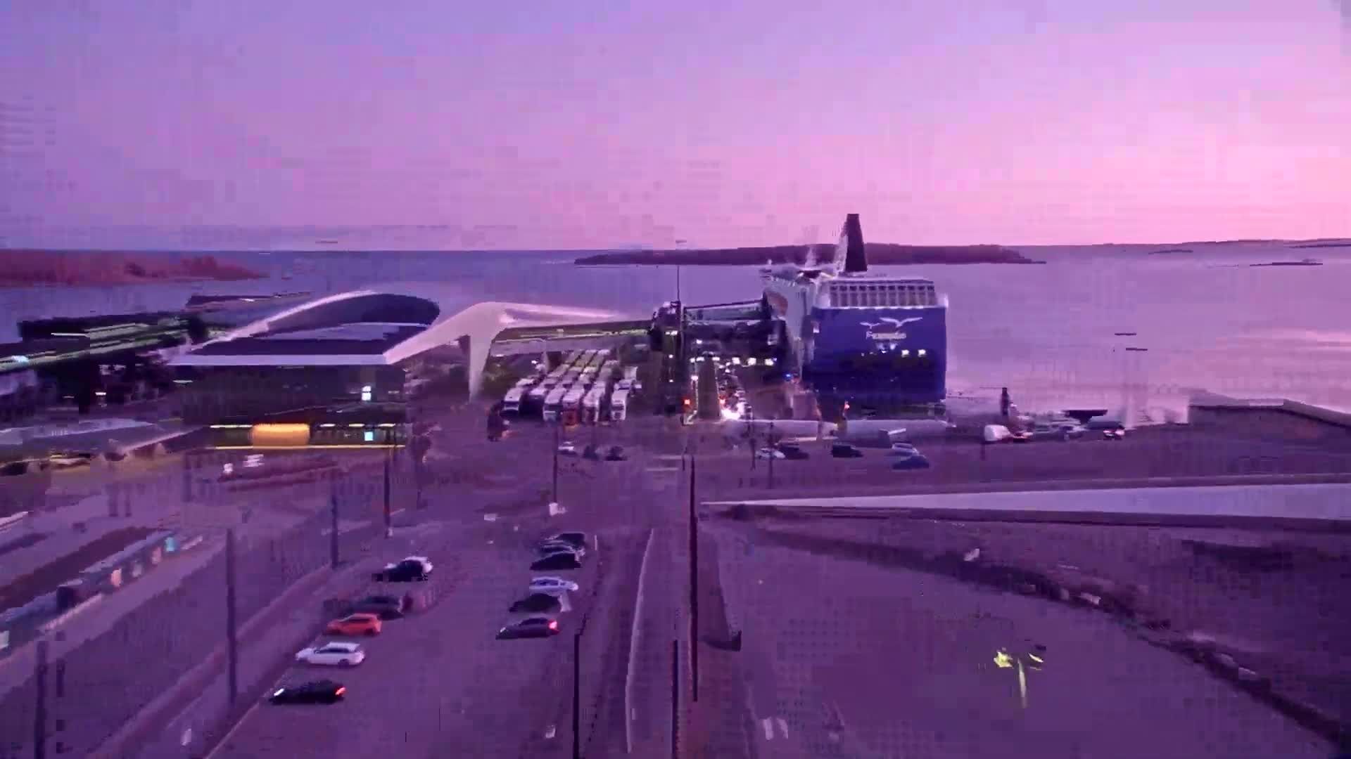 A large ferry is docked at a terminal during a purple twilight, with cars and buses surrounding the area.