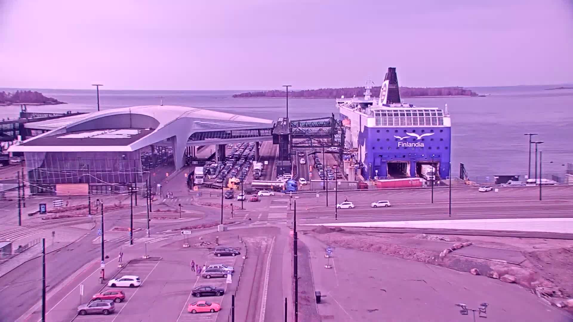 A large ferry is docked at a terminal, with numerous cars and trucks on the terminal and surrounding roads, under a pale, overcast sky.