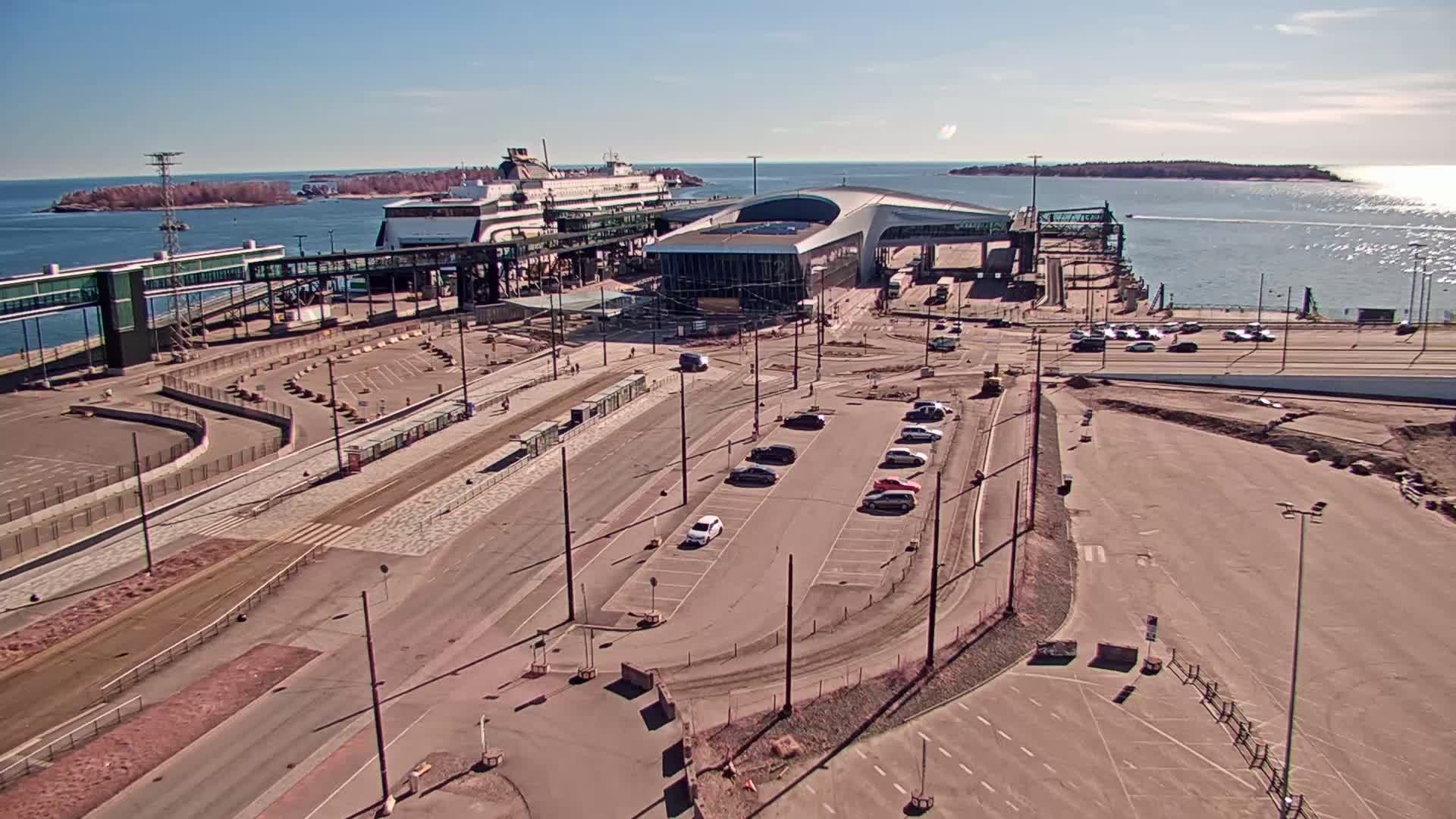 A ferry is docked at a terminal on a sunny day, with several cars parked nearby and some ongoing construction visible.
