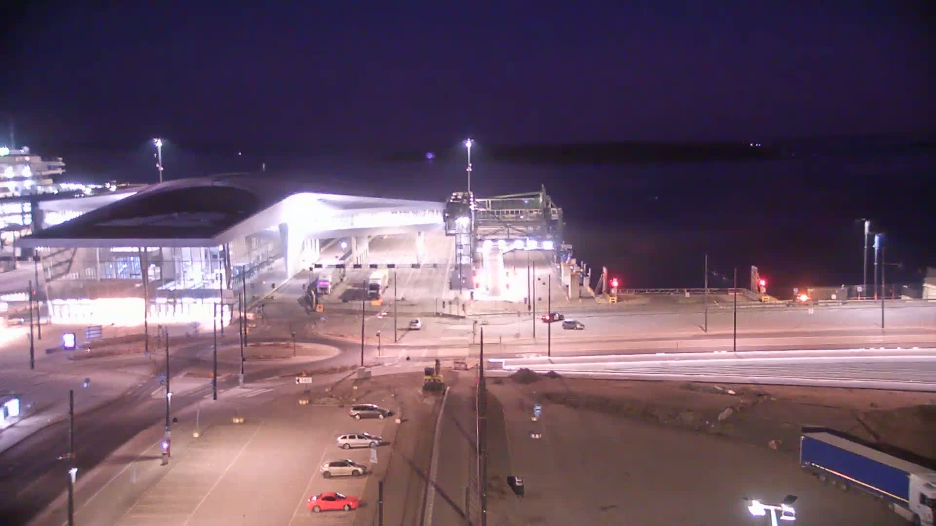 A brightly lit ferry terminal at night, with a few cars parked nearby and a dark body of water visible in the background under a clear, dark sky.