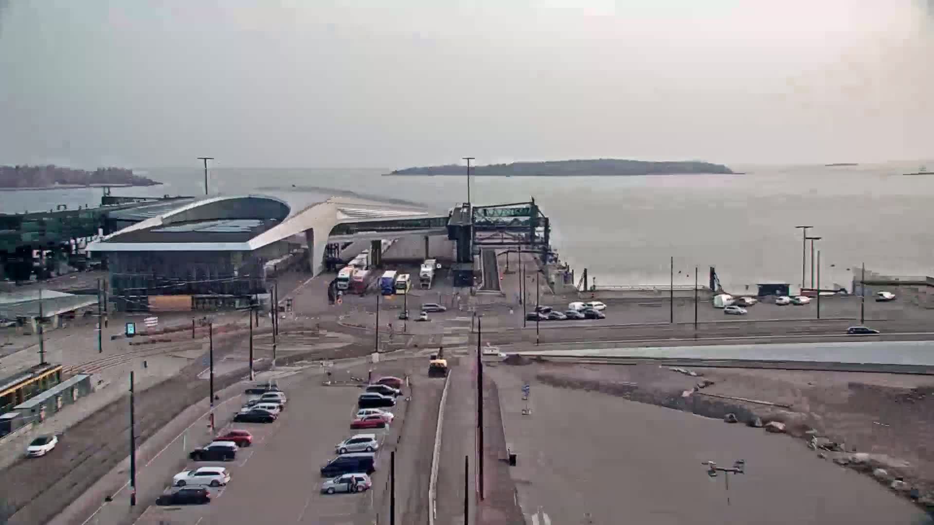 A ferry terminal with several vehicles parked nearby is shown on an overcast day, with calm water visible in the background.