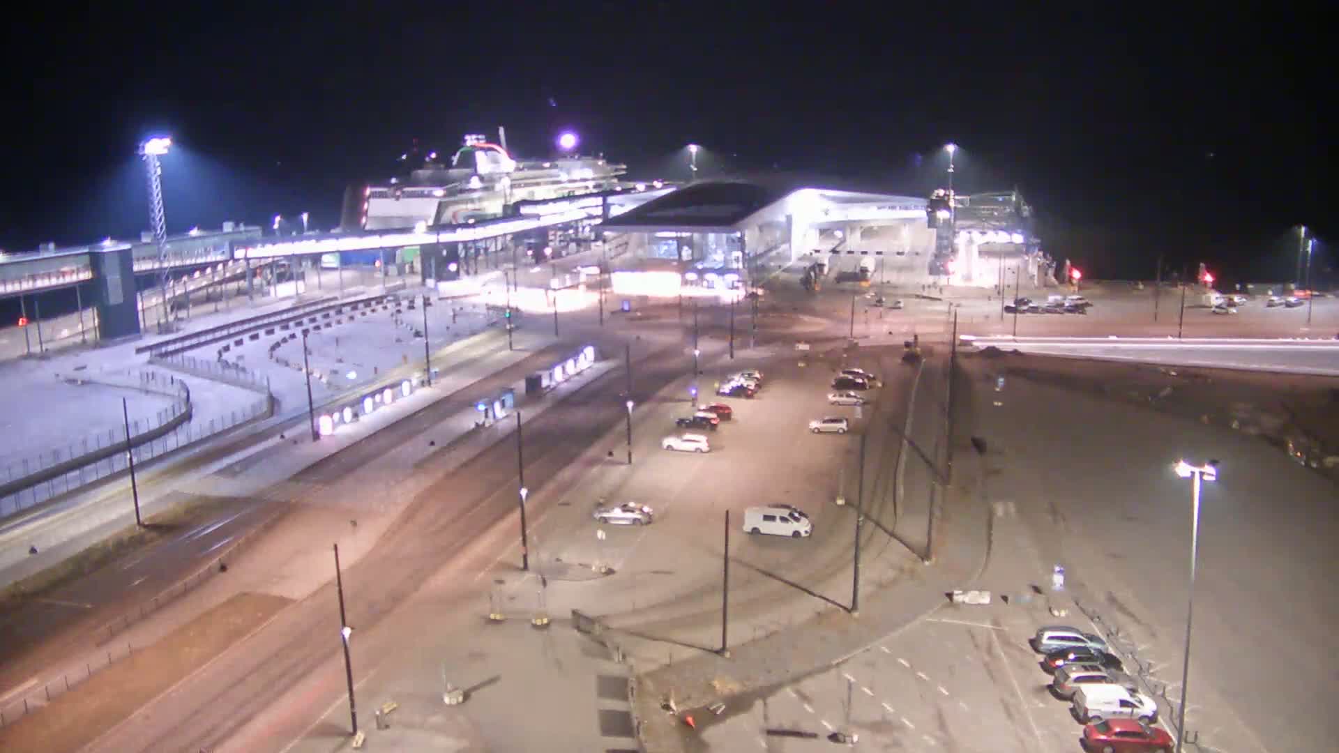 A large ferry is docked at a terminal at night, with several cars parked in lots nearby under bright lights.