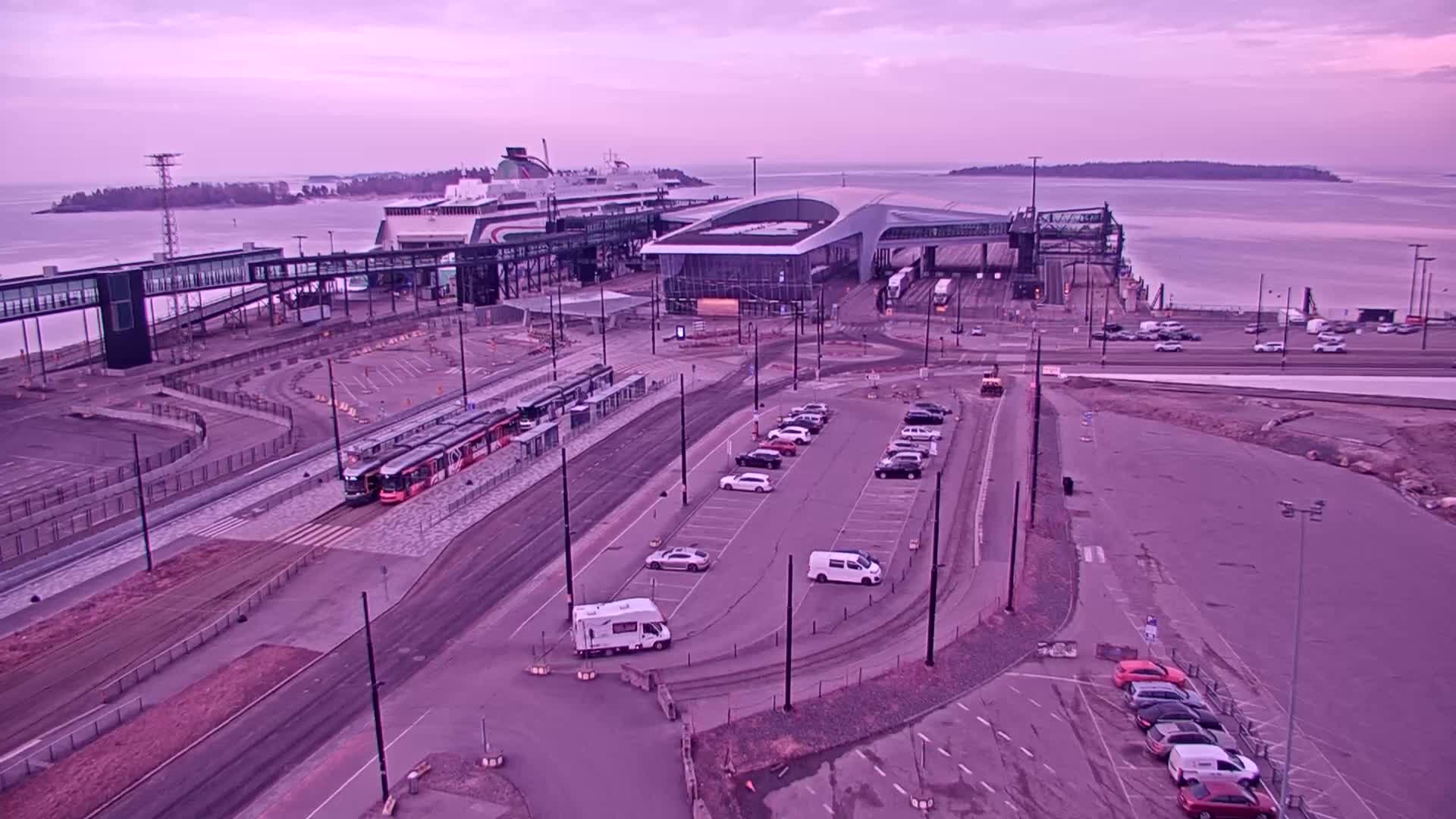 An aerial view shows a ferry terminal with trams, cars in parking lots, and a ferry docked under a purplish-pink sky.