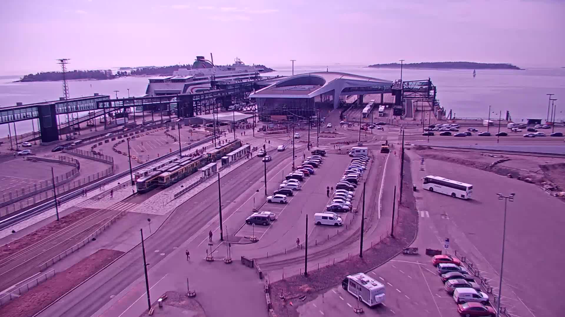 A ferry terminal on a calm, hazy day, featuring a docked ferry, a tram, several cars parked in lots, and a bus, all situated near a body of water with islands in the distance.