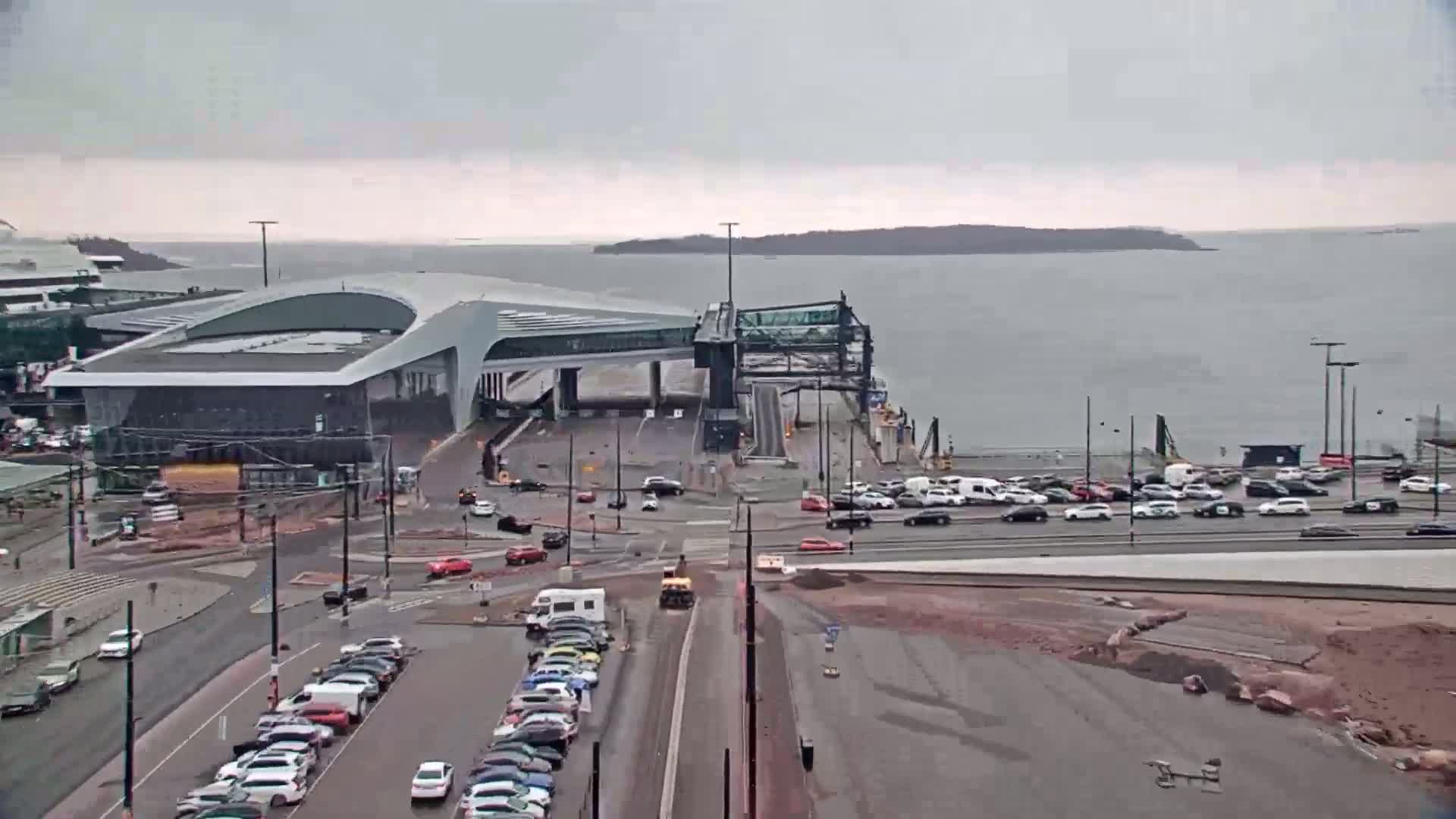 A ferry terminal with a modern building and a raised gangway is shown, surrounded by parked and moving cars on a rainy day, with calm water and a distant island visible in the background.