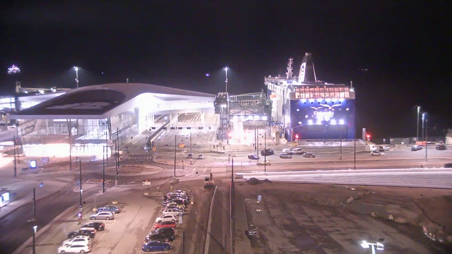 A large ferry is docked at a terminal at night, with cars parked nearby and construction or landscaping work in progress.