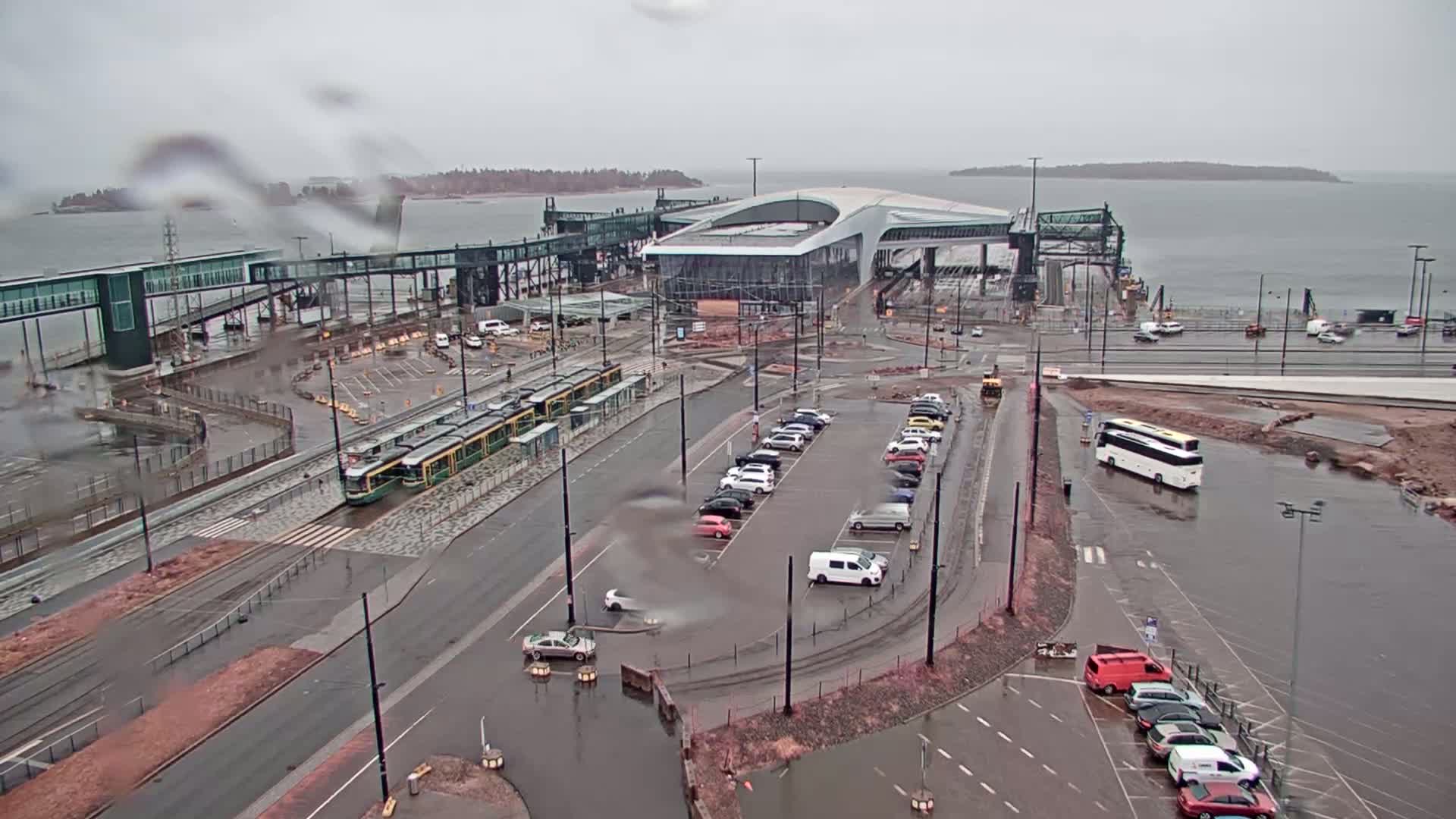 A rainy day view of a ferry terminal with trams, cars, and buses parked in a large lot, near a body of water.