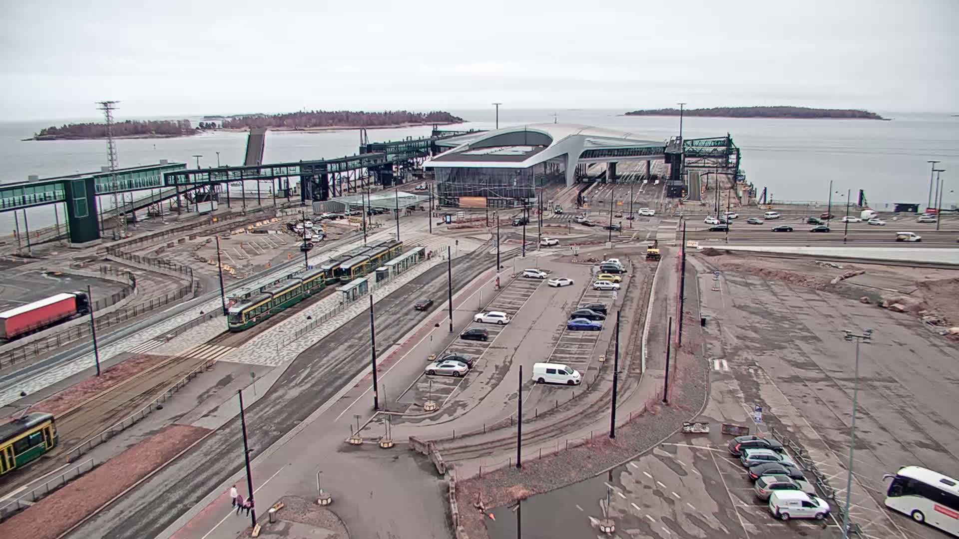 An aerial view shows a ferry terminal with trams, cars in parking lots, and a body of water under an overcast sky.