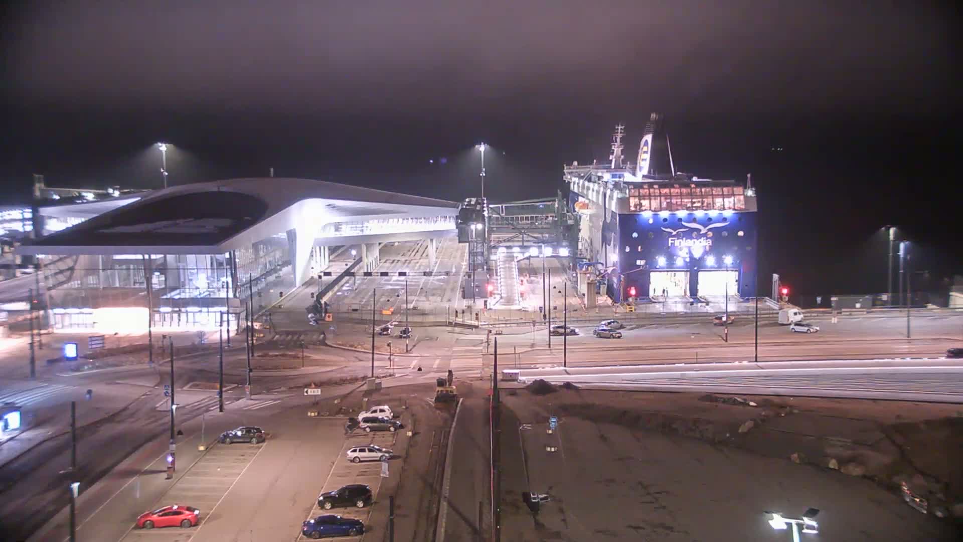 A large ferry is docked at a terminal at night under foggy conditions, with cars parked in a lot nearby.