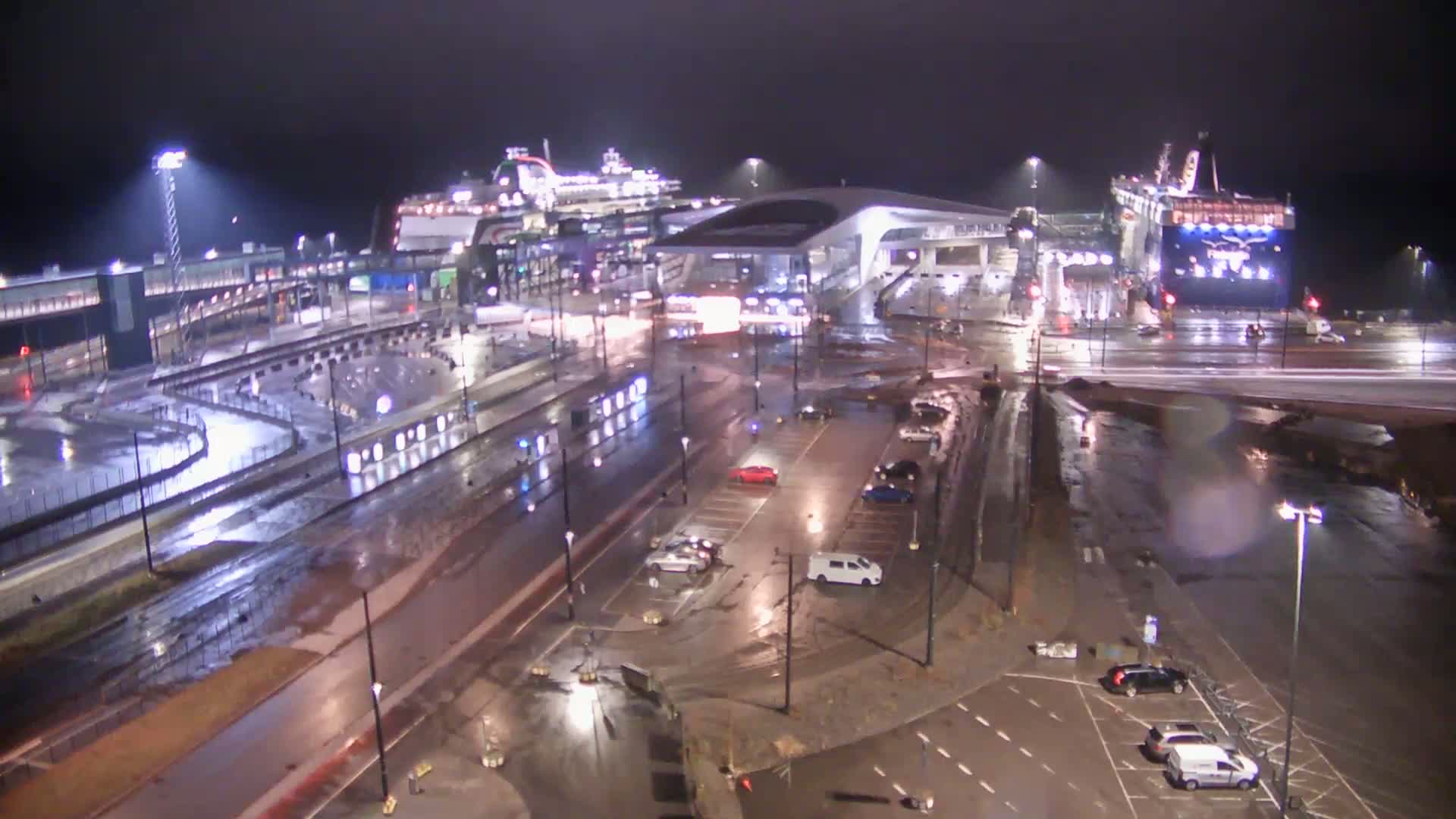 A nighttime, rain-soaked port area shows two large ferries docked near a modern terminal building, with several cars parked in adjacent lots.
