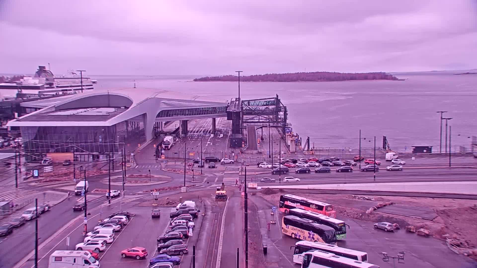 A ferry terminal with a modern building and several buses and cars parked nearby, under an overcast sky.